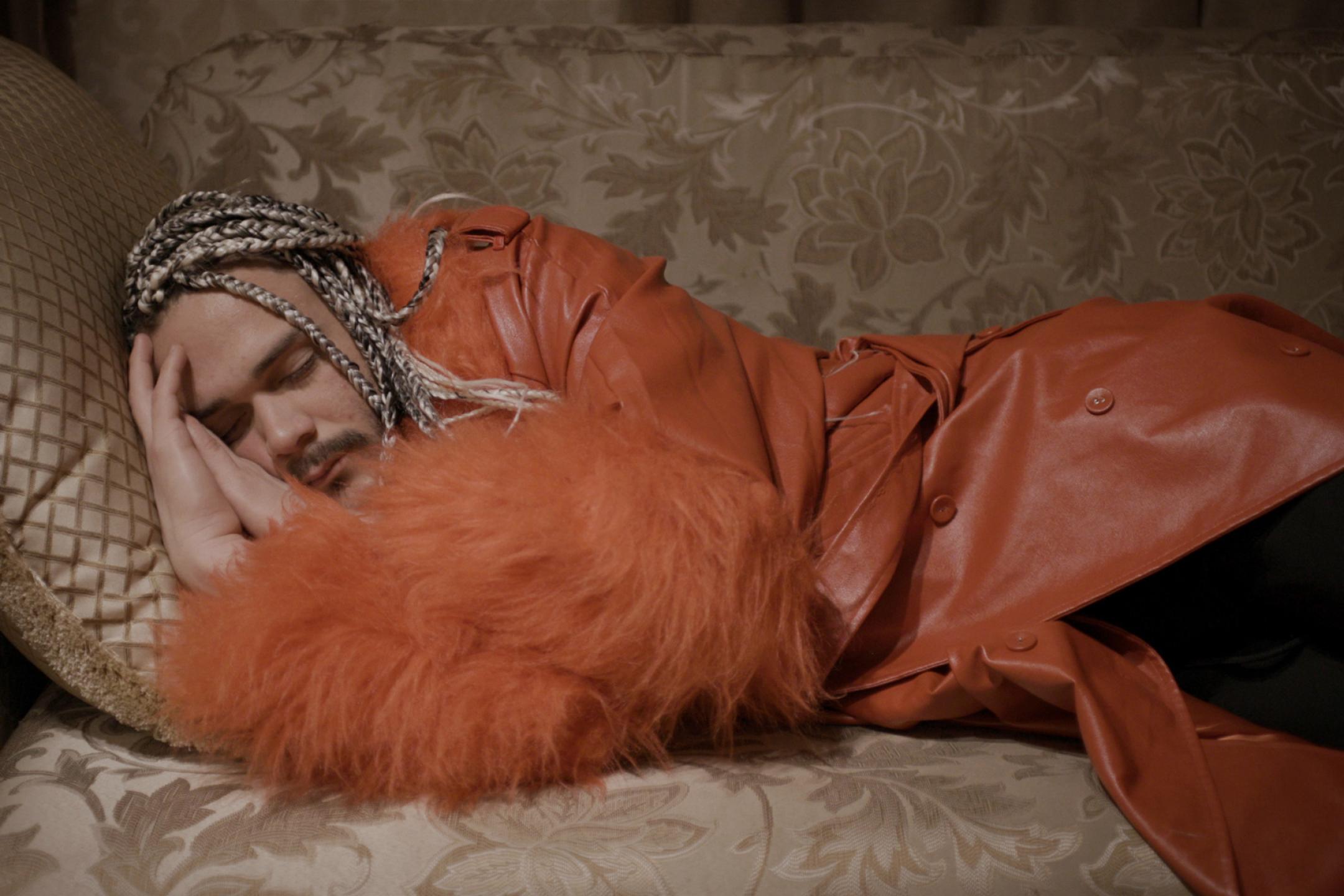 A man with blond braids and a red leather jacket lies on a sofa. He has his eyes closed.