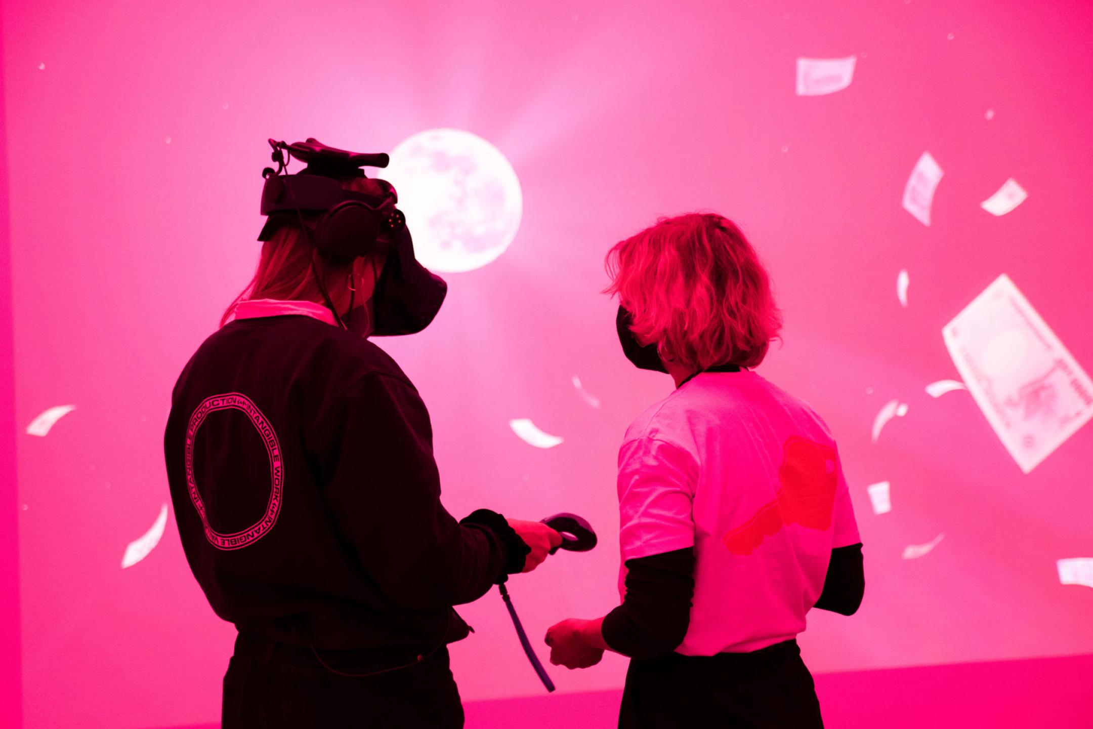 Zwei Personen stehen in einem Raum, der in Pinkes Licht getaucht ist. Vor ihnen eine riesige Leinwand, auf der ein Mond und fliegende Geldscheine zu sehen sind. Die Person links trägt eine VR-Brille.