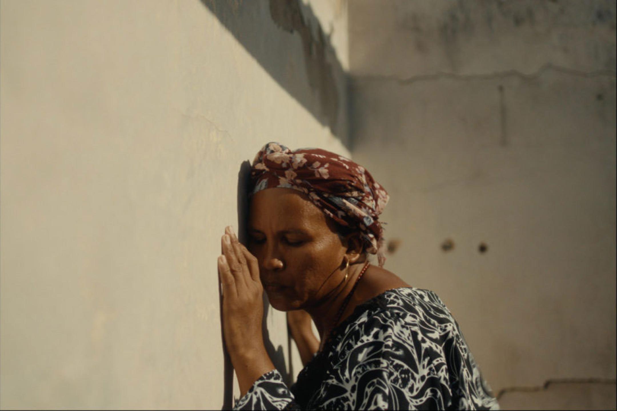 A woman leans with her head and hands against a wall in the sun.