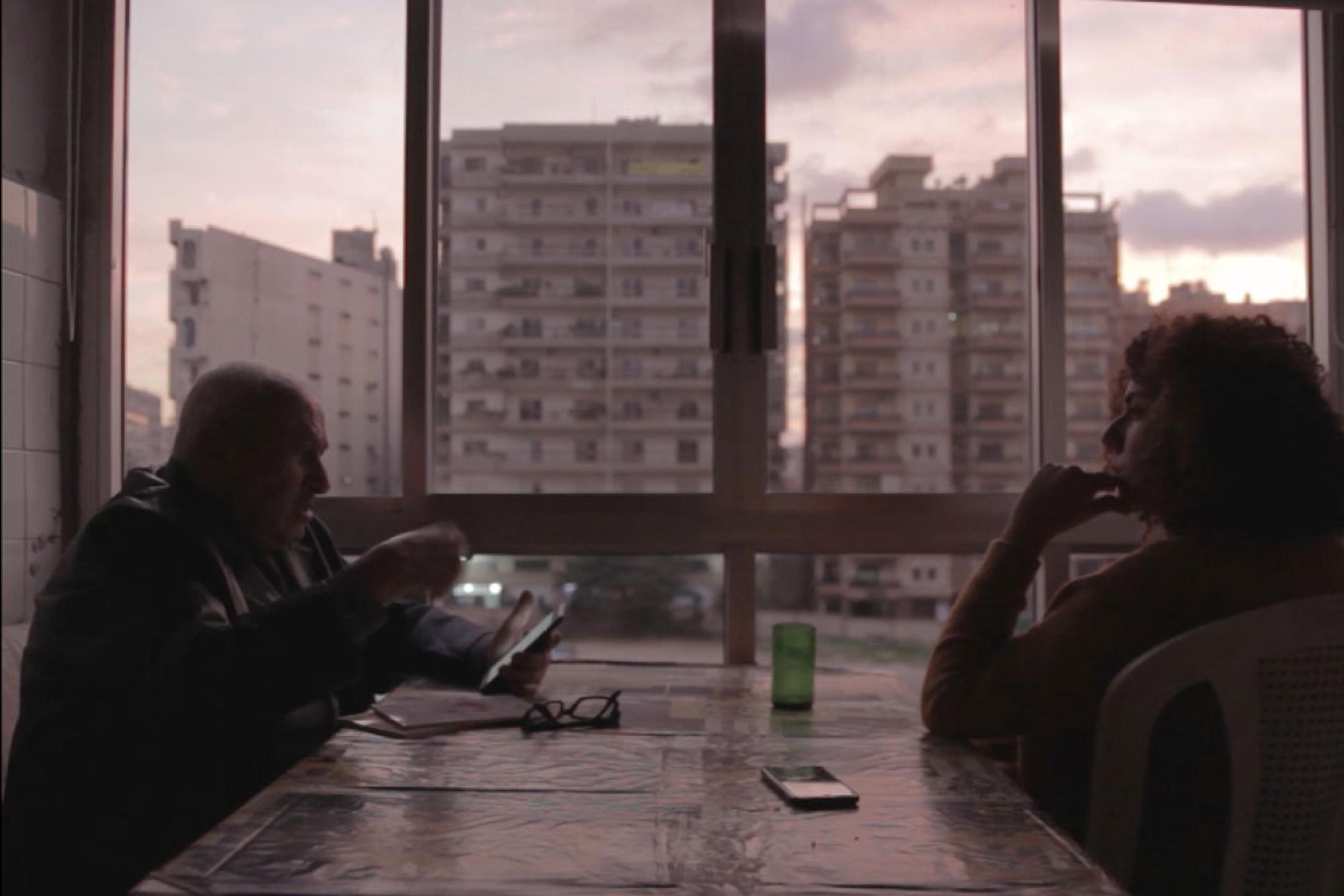 A man and a woman sit opposite to each other at a table at a window. The view shows several residential blocks.