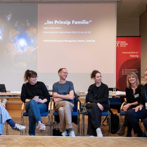 In einem Tagungsraum sitzen sechs Menschen gut gelaunt nebeneinander. Ein Mann ganz rechts hebt seinen Arm einladend in Richtung Publikum, hinter Ihnen zeigt eine Leinwand den Text "Im Prinzip Familie" und ein Filmstill mit mehreren Kindern, die ein Feuerwerk anzünden.