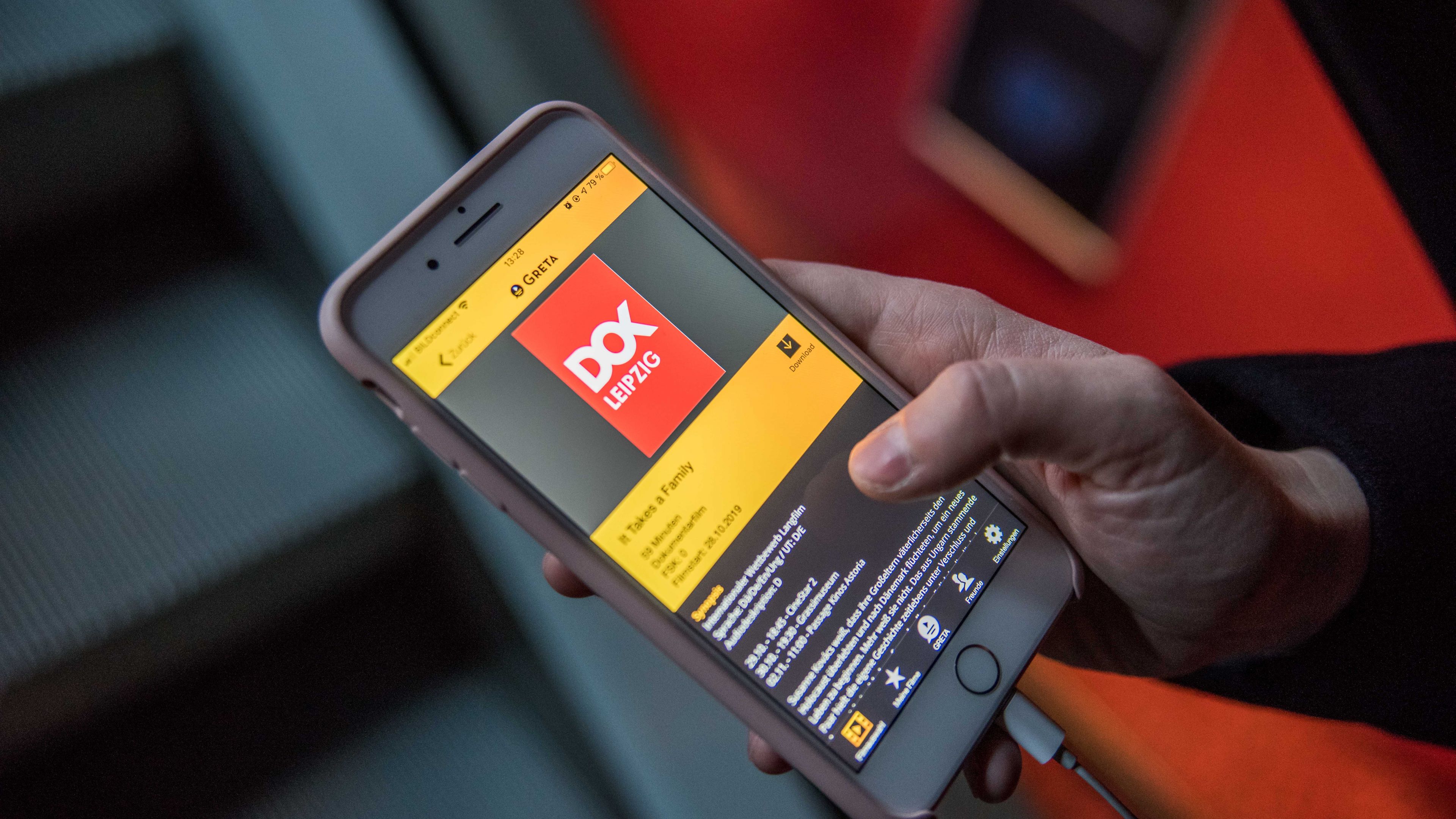 Eine Besucherin, von der nur die rechte Jand zu sehen ist, hält darin ihr Smartphone. Auf dem Display erscheint die App Greta, ausgewählt ist der Film It Takes a Family.