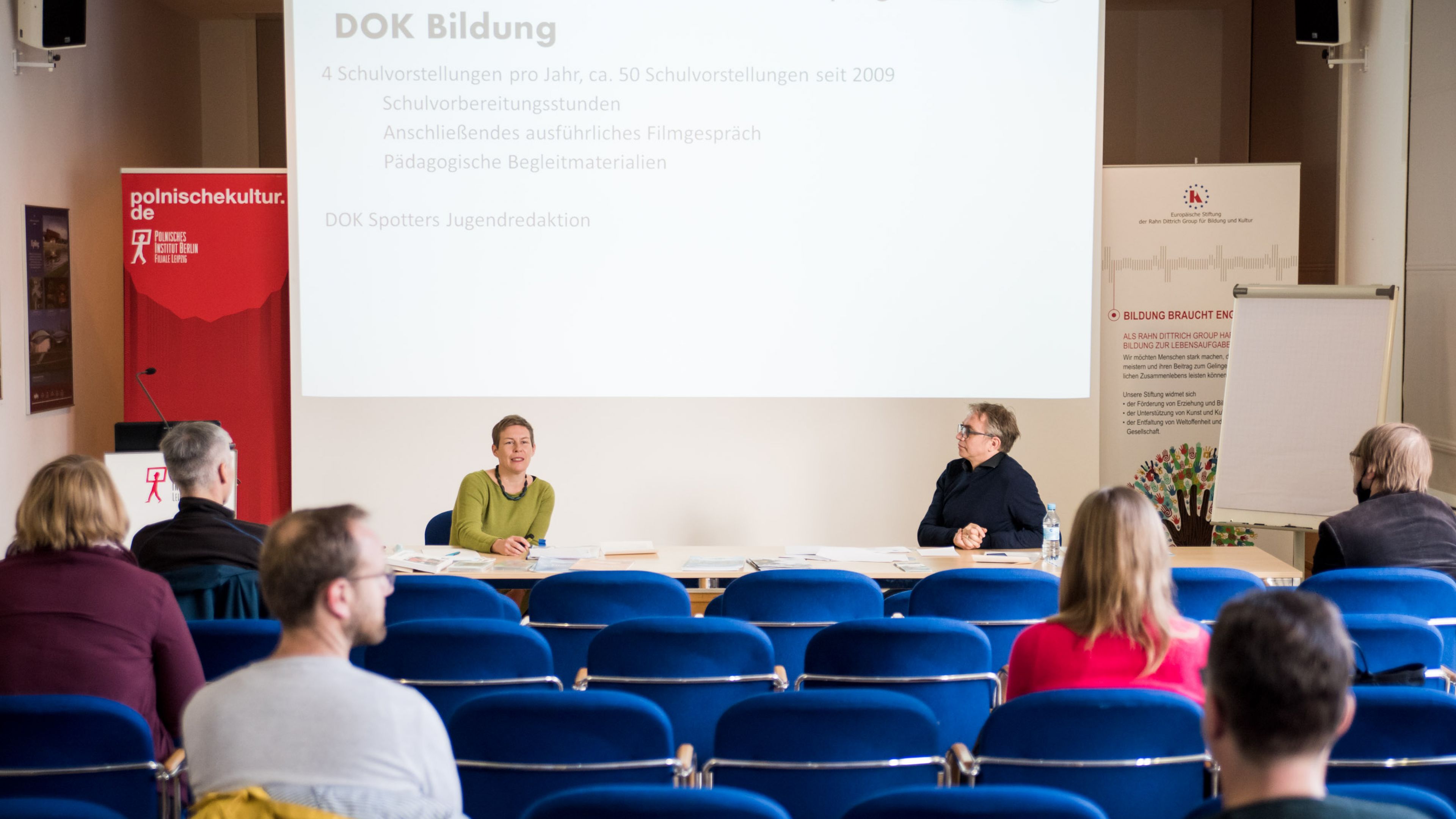 Ein Konferenzraum mit einer Frau und einem Mann, die auf dem Podium sitzen und zum Publikum sprechen, hinter ihnen eine Leinwand.