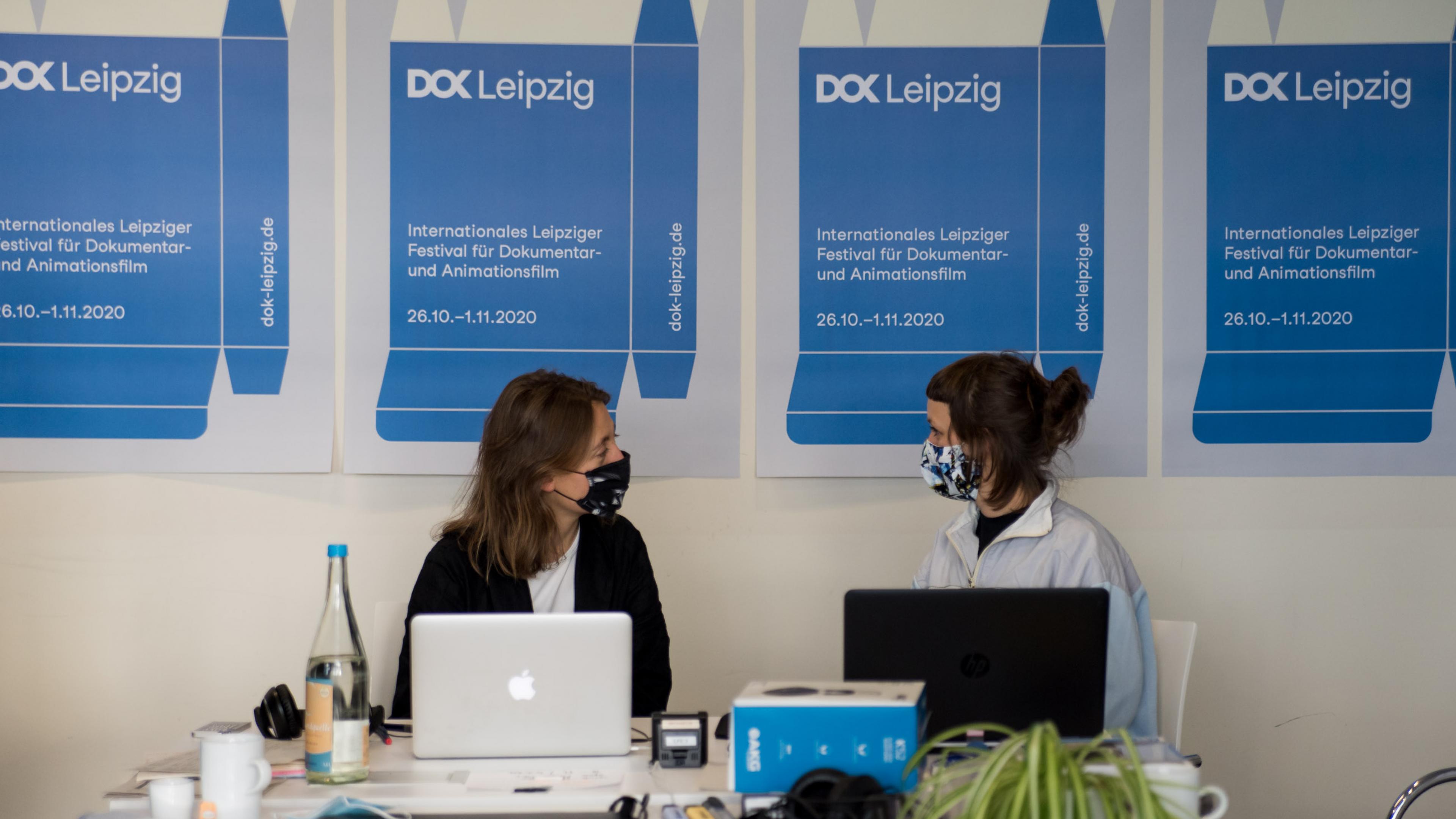 A row of DOK Industry posters on the wall, in front of them two women with masks are sitting next to each other in fron of their laptops and look at each other