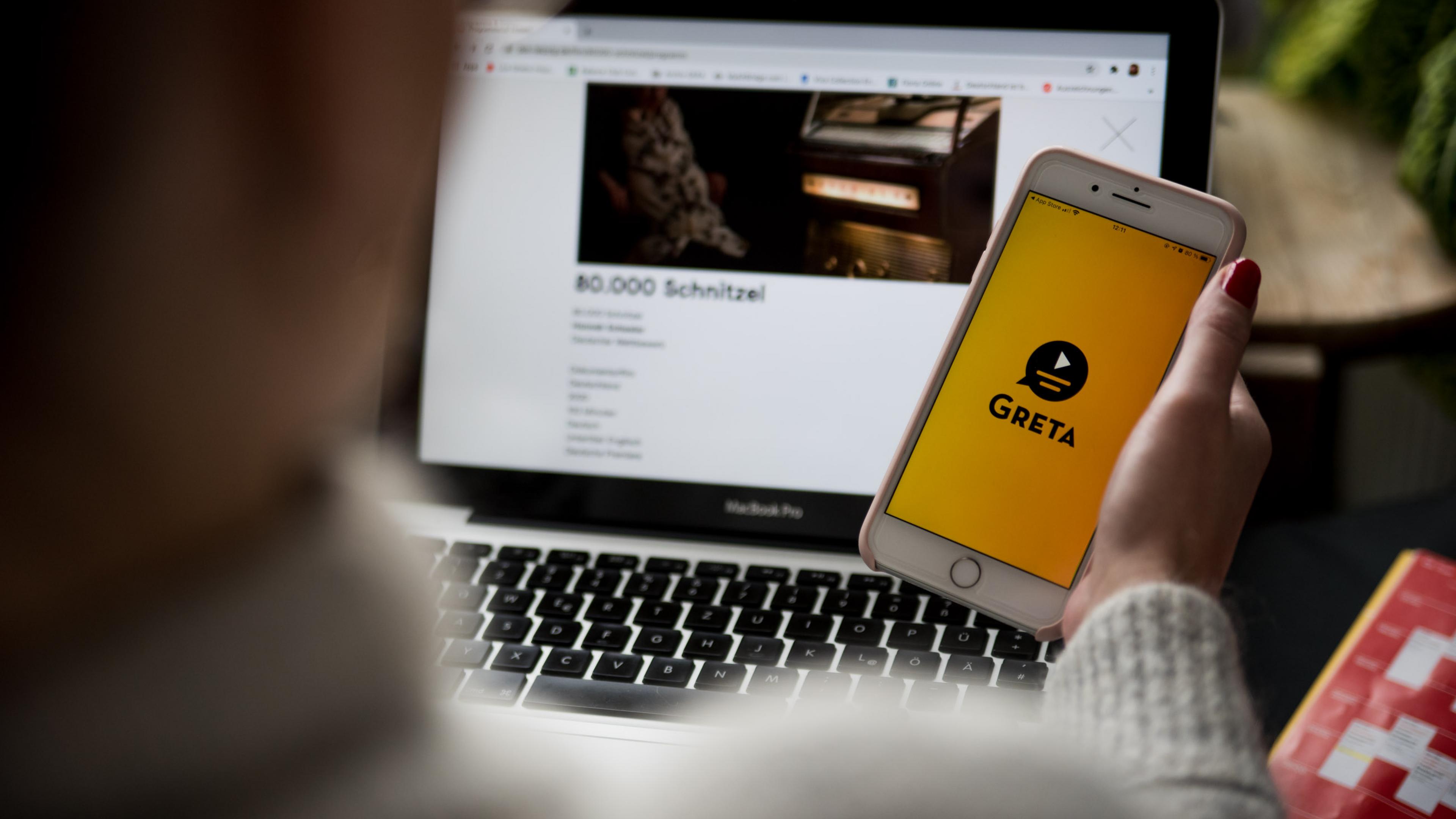 Blick über die Schulter einer Person auf ihren Laptopbildschirm, in der Hand hält sie ein Smartphone mit dem Startbildschirm der App Greta.