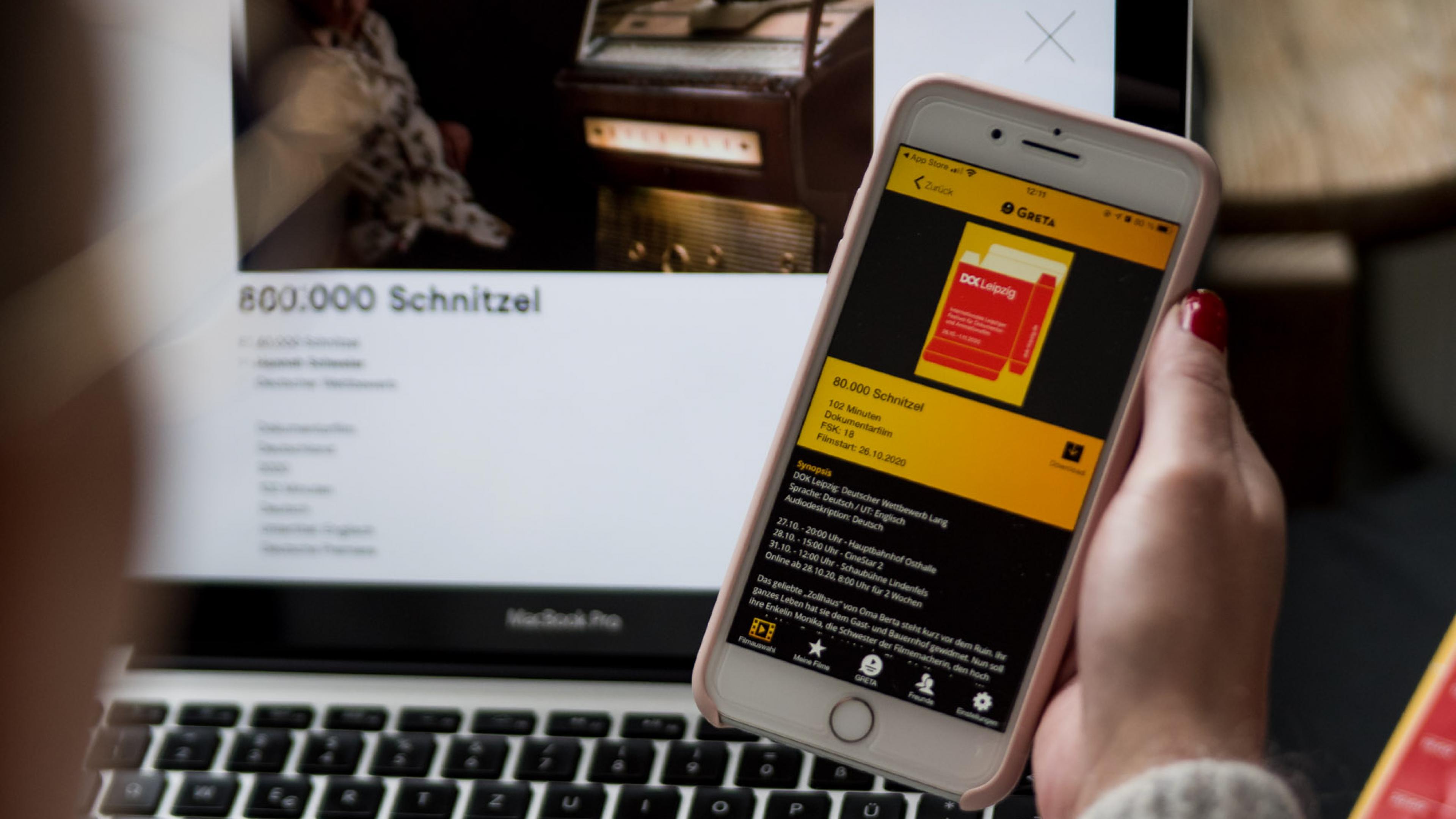 Blick über die Schulter einer Person auf ihren Laptopbildschirm, in der Hand hält sie ein Smartphone mit der App Greta.