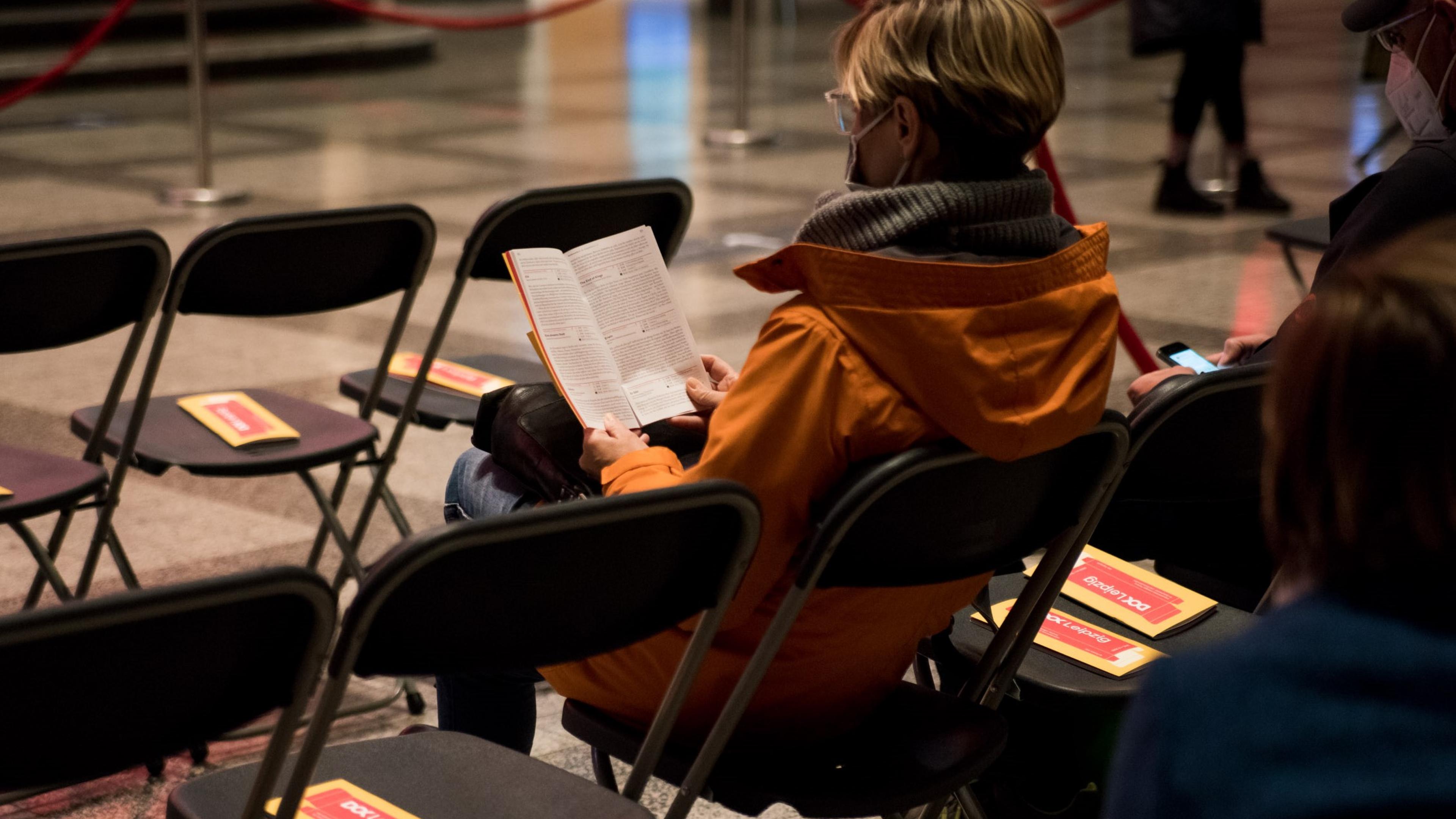 Eine Frau mit Maske sitzt auf einem Stuhl und liest in einem DOK Leipzig Programmheft. Um sie herum stehen mehrere Stühle in Reihen mit Programmheften auf den Sitzen, neben ihr sitzt ein Mann mit Maske