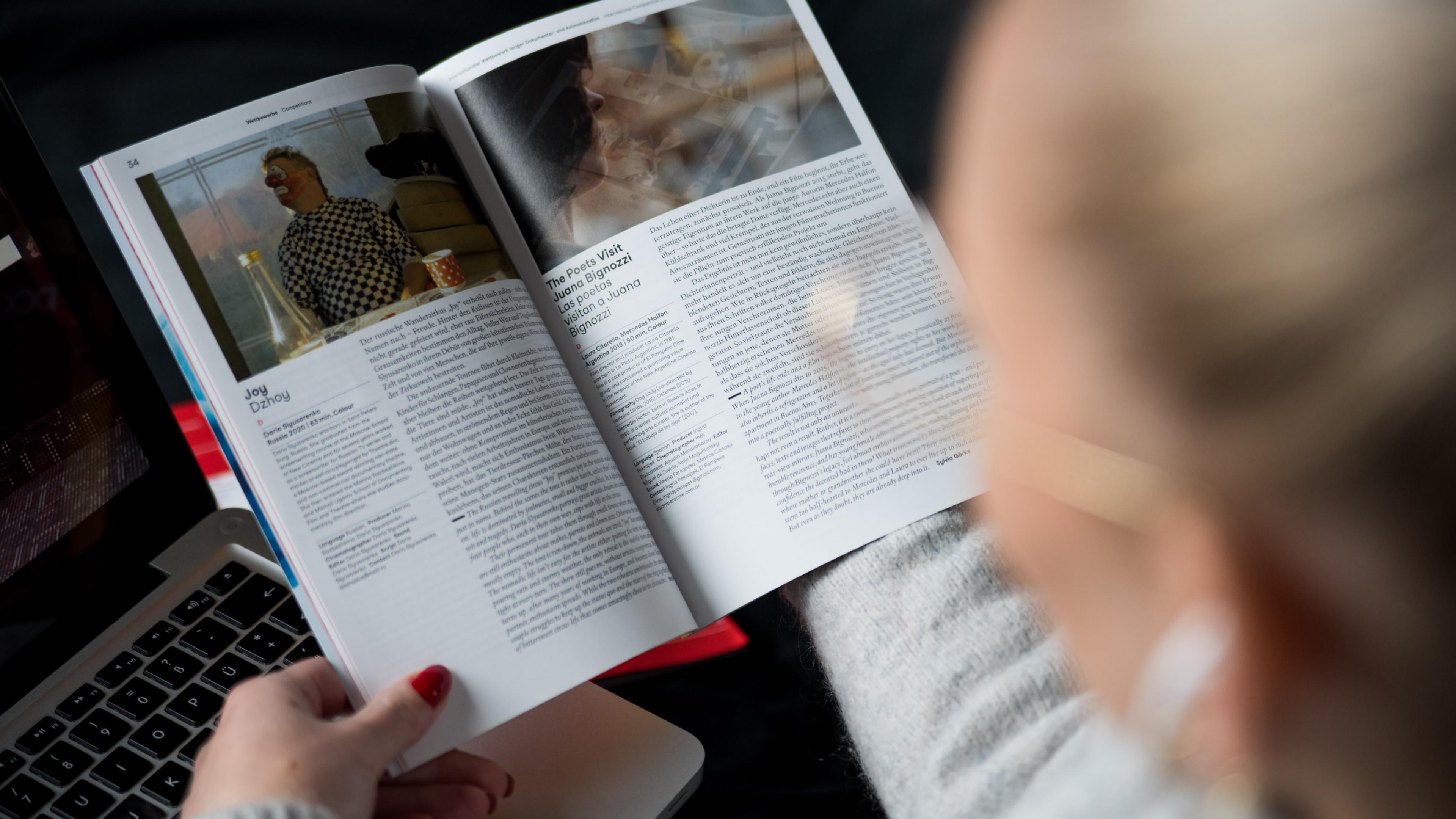 Eine Frau blättert im DOK Festivalkatalog von 2021, die Kameraperspektive blickt über ihre Schulter auf den Katalog in ihrer Hand.