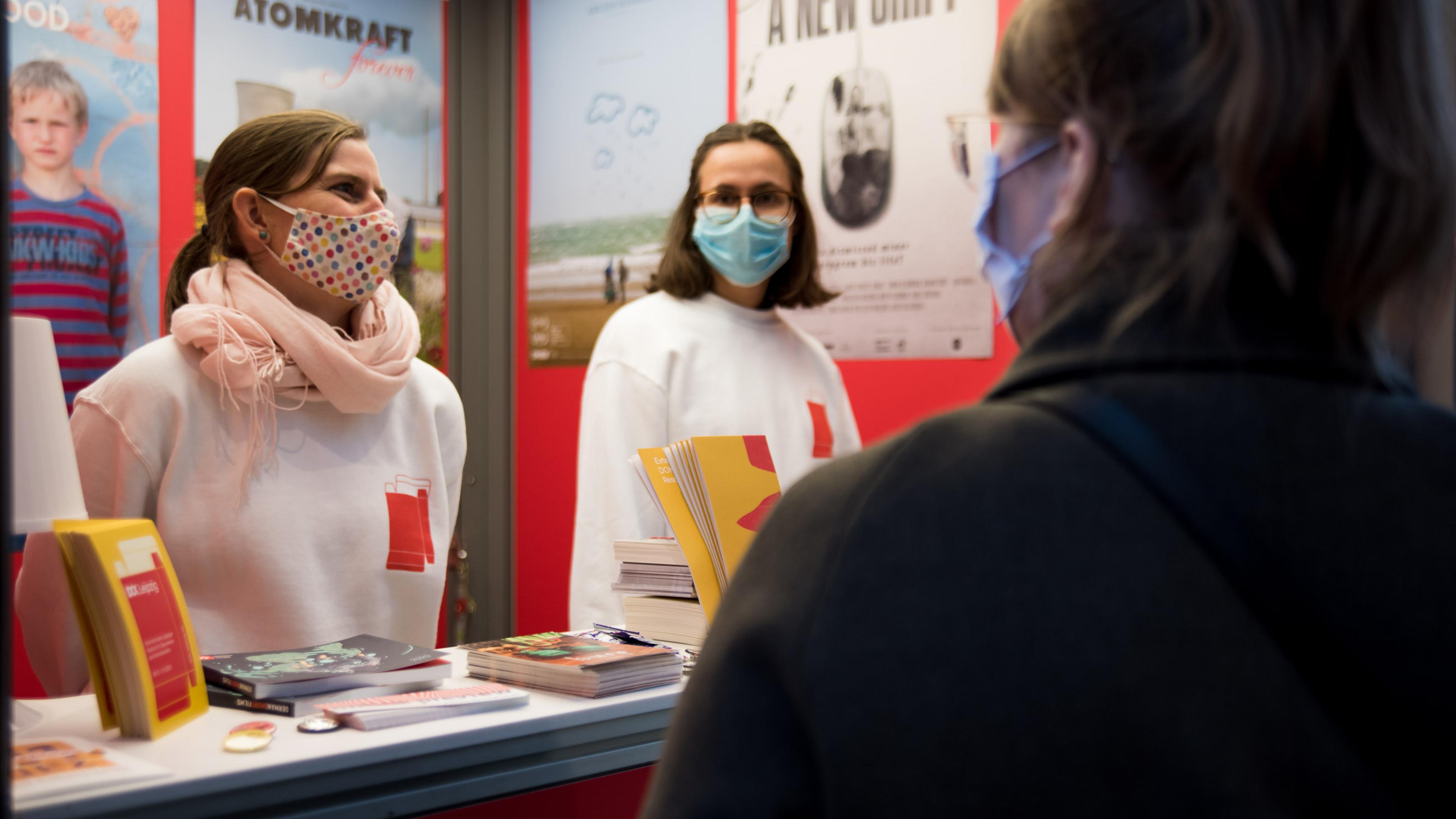 Zwei Volunteers mit DOK Pullis im Gespräch mit einer Besucherin am Info-Schalter.