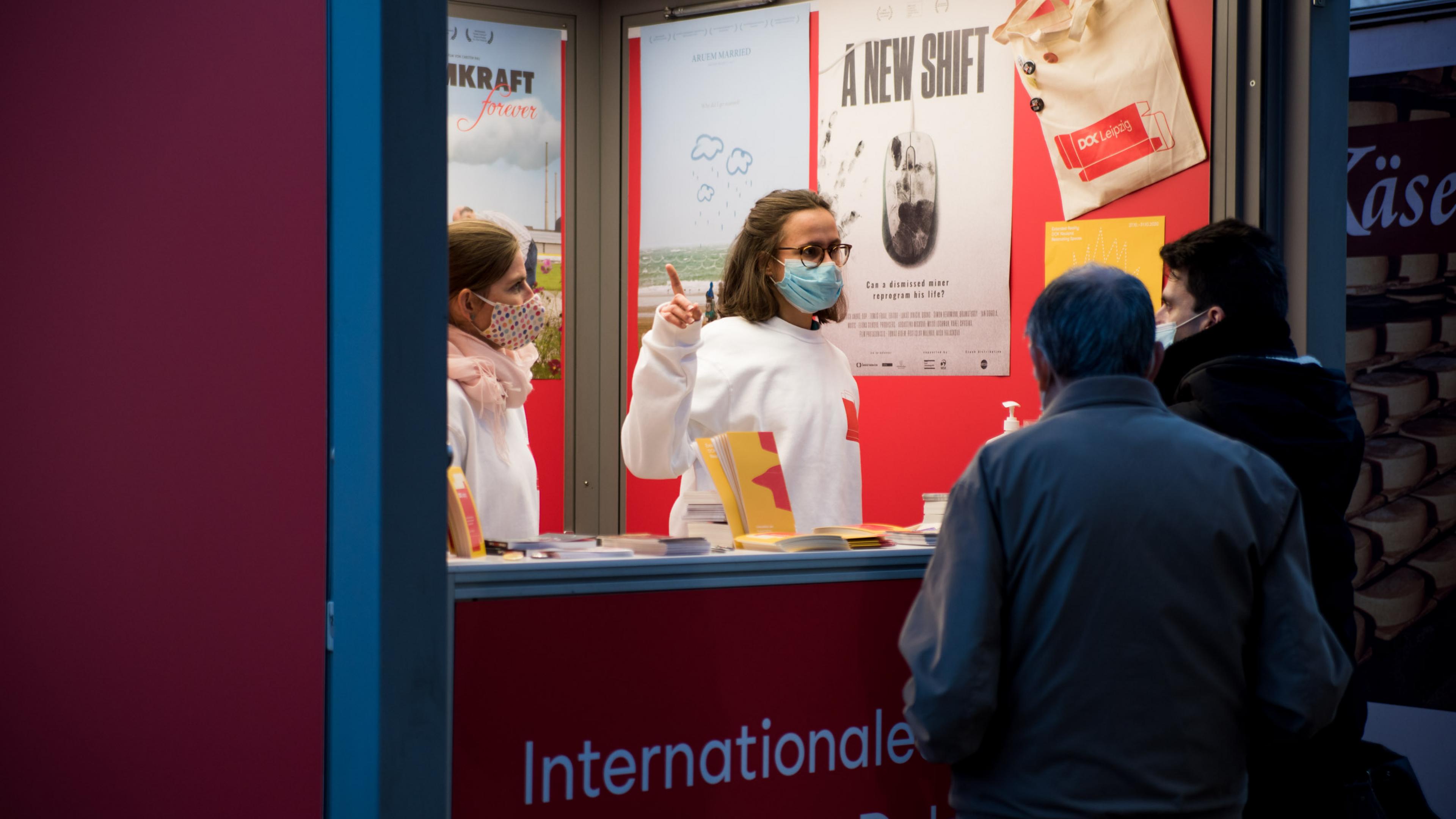 Zwei DOK-Mitarbeiterinnen stehen nebeneinander am Infostand. Eine von ihnen spricht zu zwei Gästen und zeigt mit der Hand nach rechts.