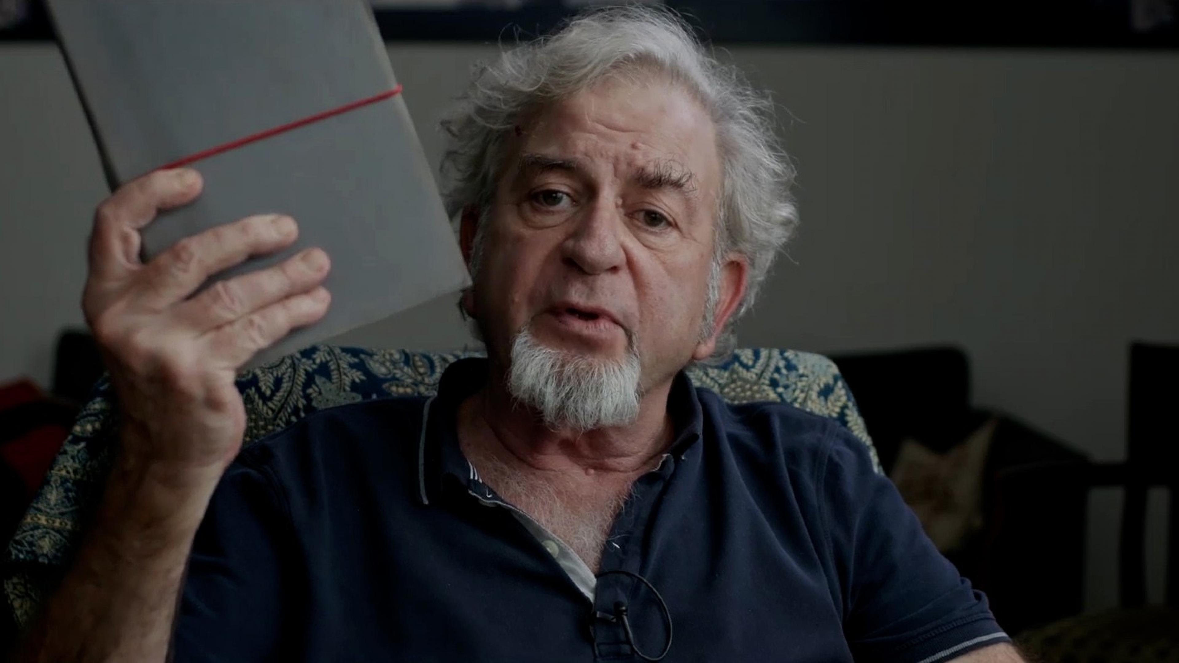 Regisseur Avi Mograbi blickt in die Kamera, spricht dabei und hält dabei in der rechten Hand ein Notizbuch hoch.