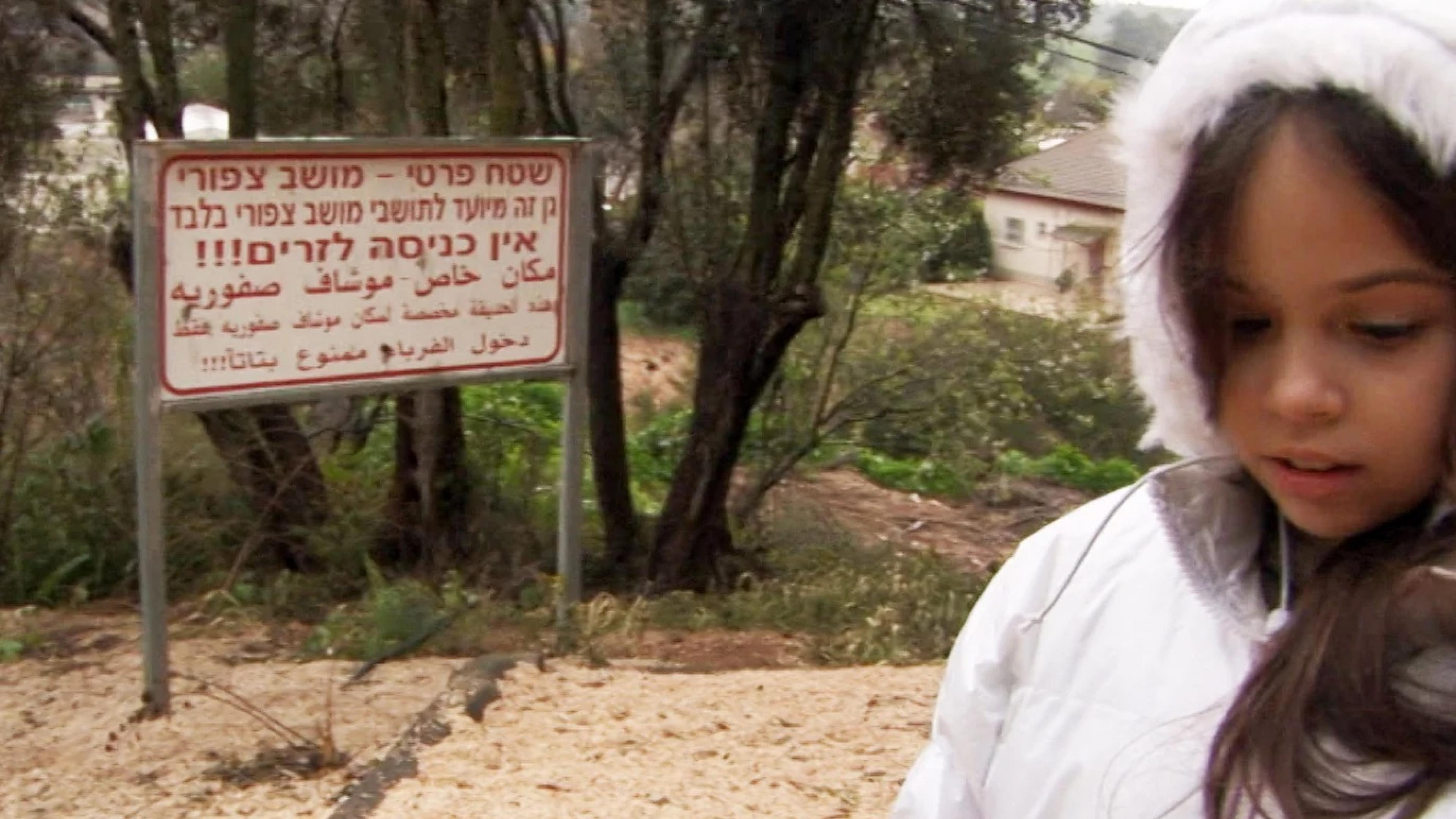 Filmszene aus "Once I entered the Garden": Ein Mädchen in einer Winterjacke blickt auf den Boden, neben ihm steht ein Hinweisschild in hebräischer und arabischer Schrift.