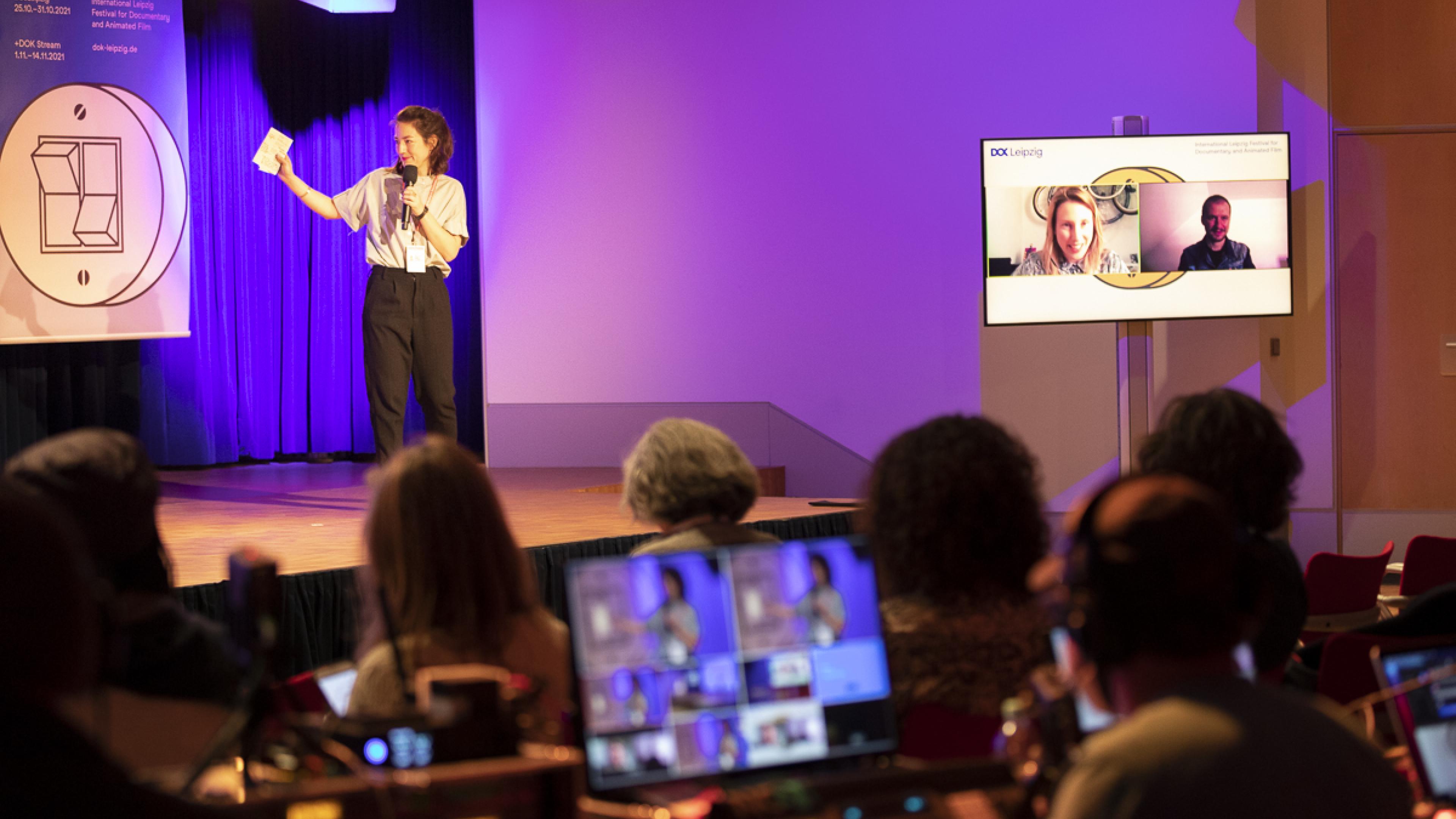Laura Klöckner is moderating a panel on stage, at her right a huge screen with two participants in Zoom, in front several screens of an editing suite.