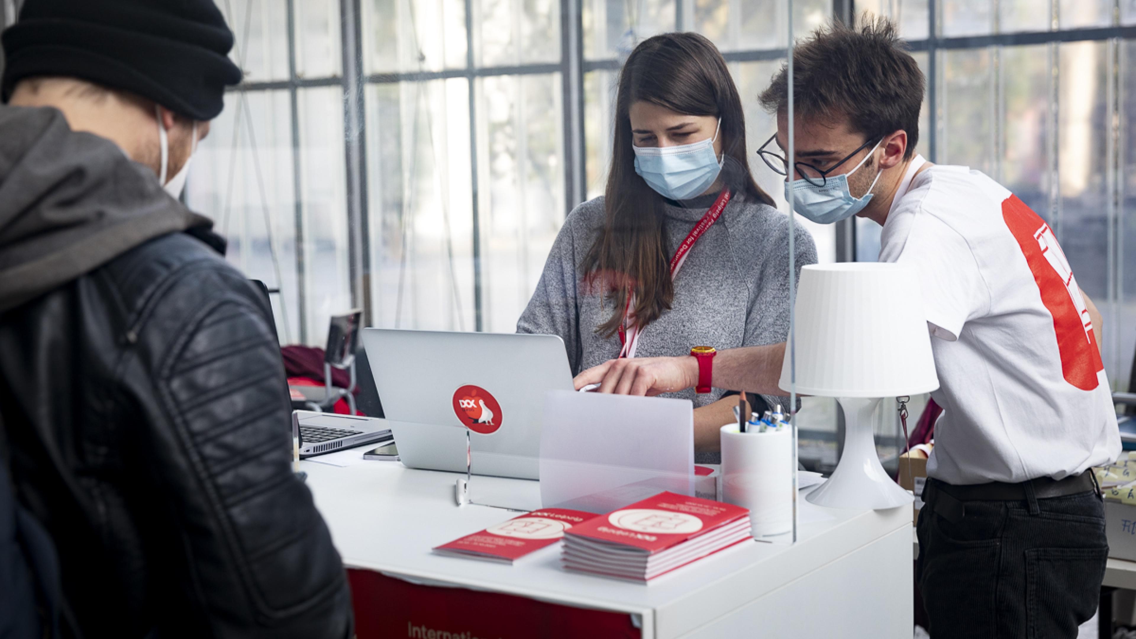 Zwei DOK-Mitarbeiter*innen beugen sich über einen Computerbildschirm am Gästecounter des Festivals. Ihnen gegenüber auf der anderen Seite des Counters steht ein Besucher.