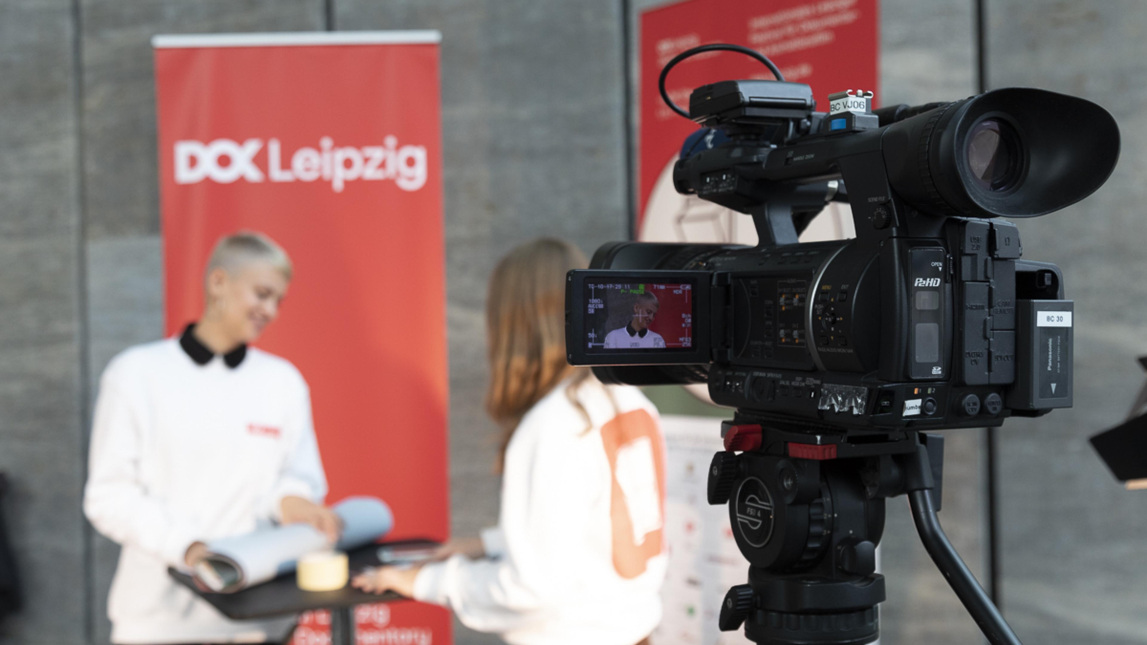 Zwei DOK Spotters proben eine Interviewsituation an einem Stehtisch. Rechts von ihnen steht eine semi-professionelle TV-Kamera auf einem Stativ. Im Hintergrund sind Aussteller mit dem Festivalmotiv.