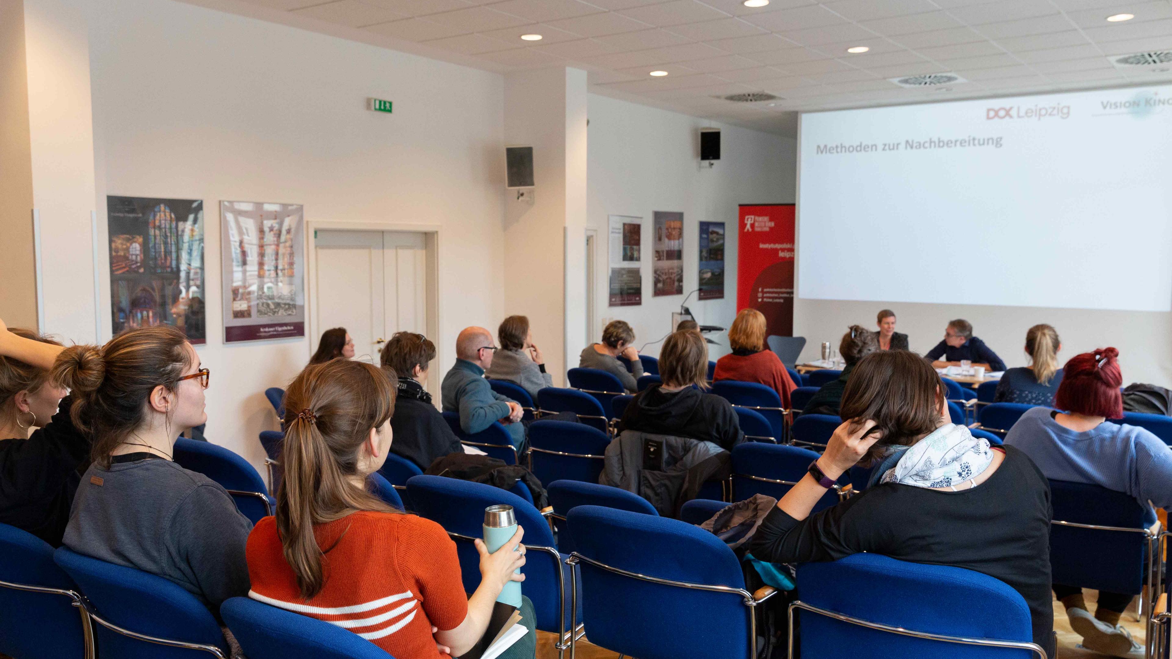 Ein Veranstaltungsraum, in dem verteilt mit Mindestabstand mehrere Personen sitzen und drei Vortragenden zuhören. Auf der Leinwand hinter den Vortragenden steht der Schriftzug: Methoden der Nachbereitung.