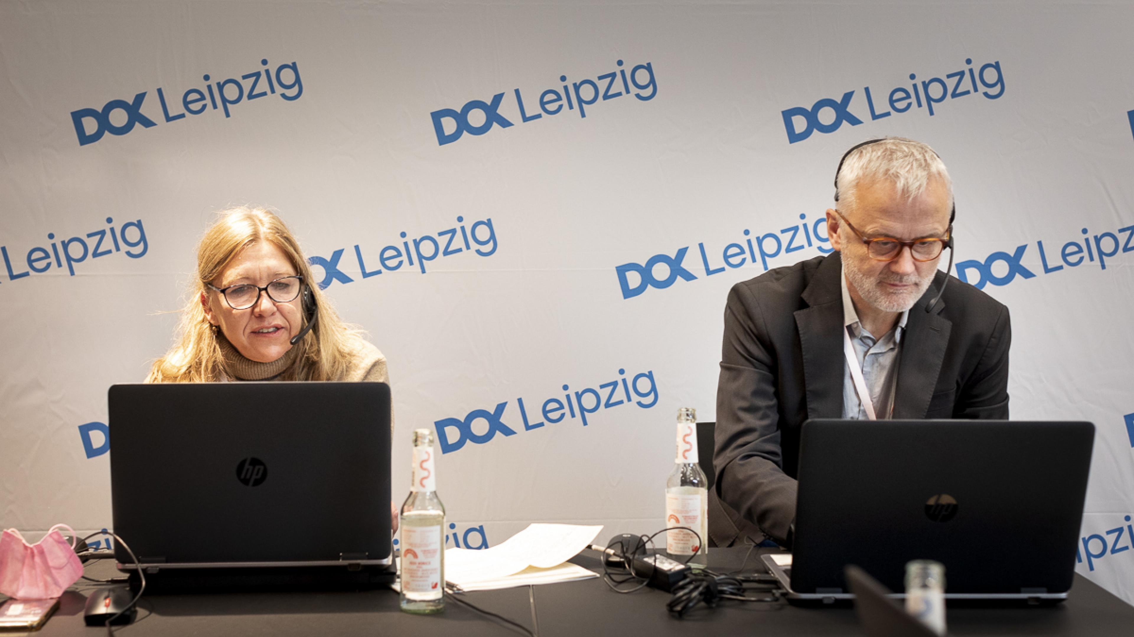 Nadja Tennstedt und Christoph Terhechte sitzen nebeneinander an zwei Laptops und blicken konzentriert auf die Bildschirme. Hinter ihnen eine Logowand mit dem DOK Leipzig Logo in blau.