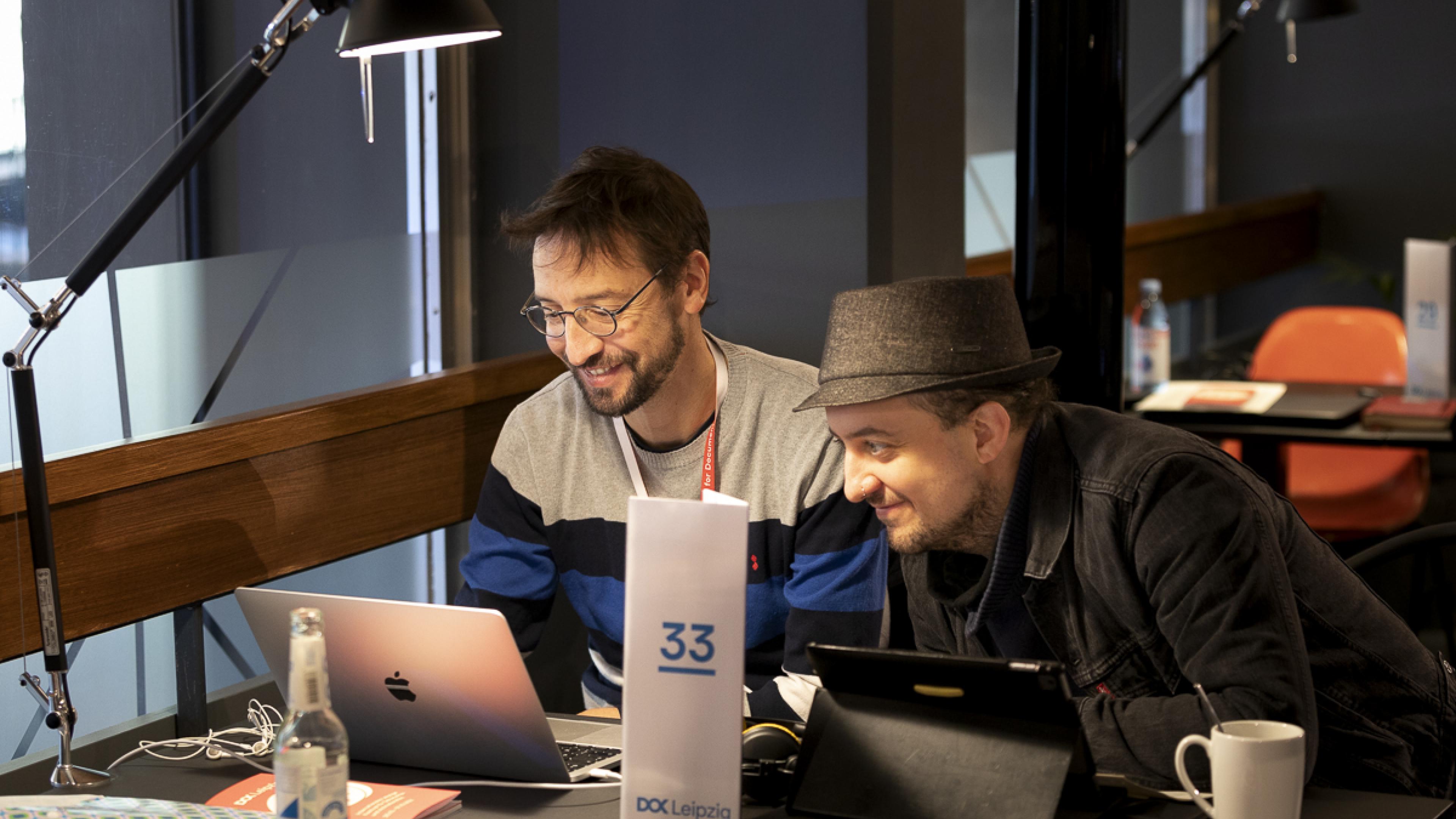 Two male participants of the Co-Pro Market sitting next to each other at a desk, looking at one laptop display. Both are laughing.
