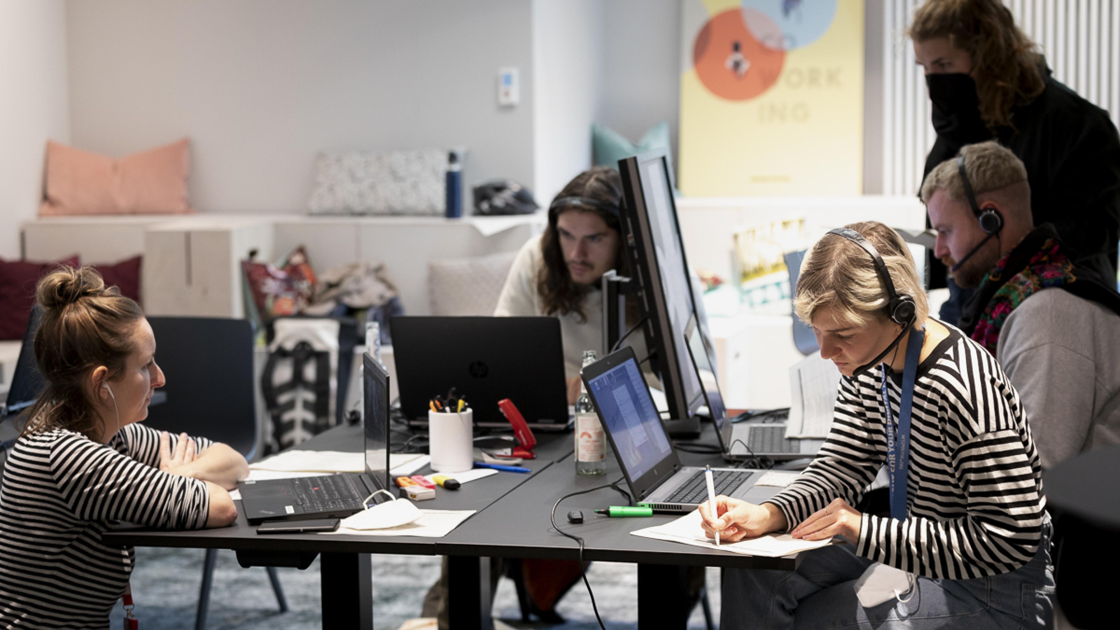 Fünf Personen gruppieren sich sitzend und stehend um einen großen quadratischen Arbeitstisch mit mehreren Laptops. Eine Frau (rechts im Bild) macht sich Notizen auf einen DIN A4-Zettel.