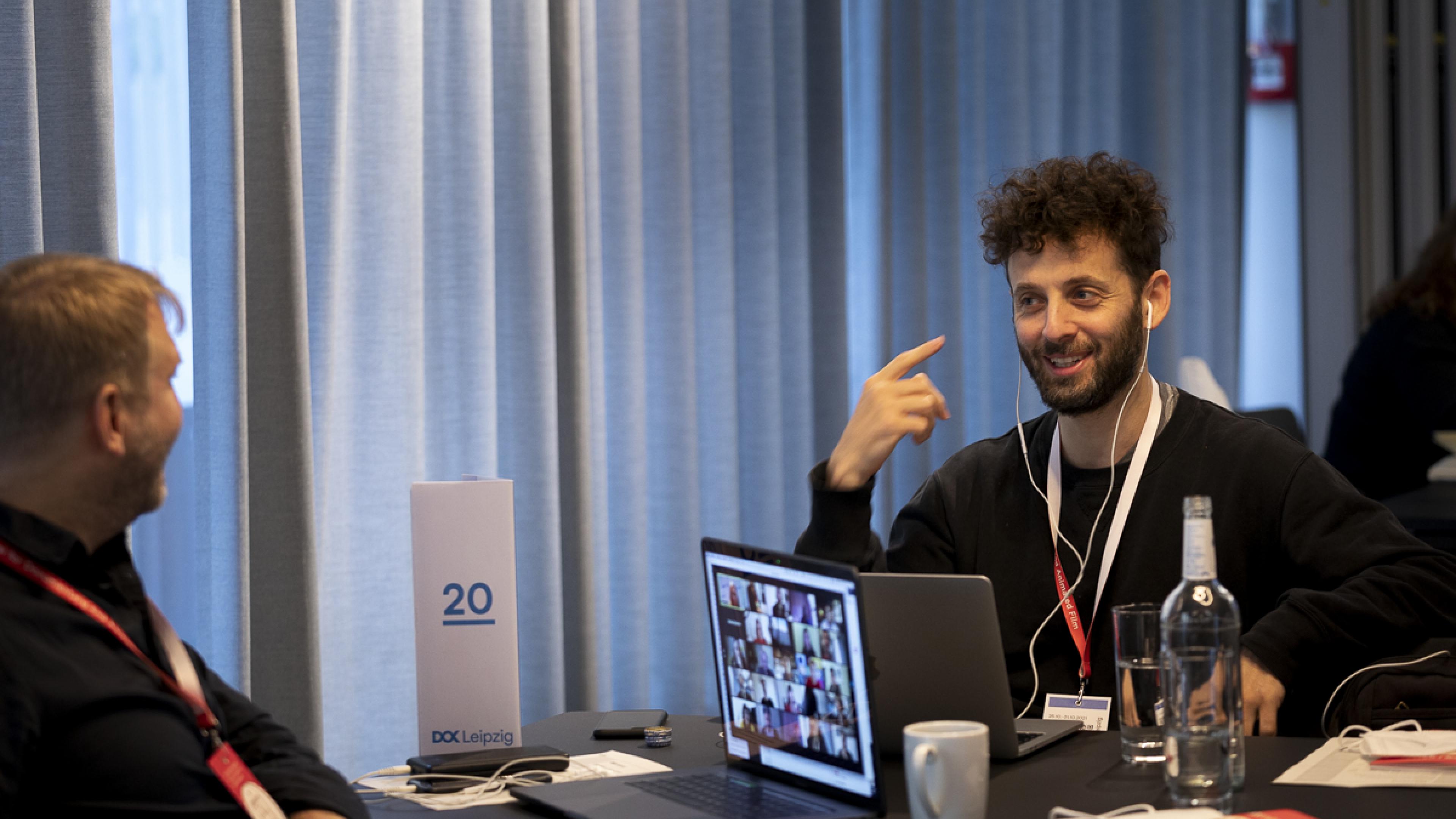 Two men are sitting opposite to each other at a desk. The men at the right smiles and points with his hand to the earphones he has plugged in. The one at the left is leaning back in his chair, smiling as well.