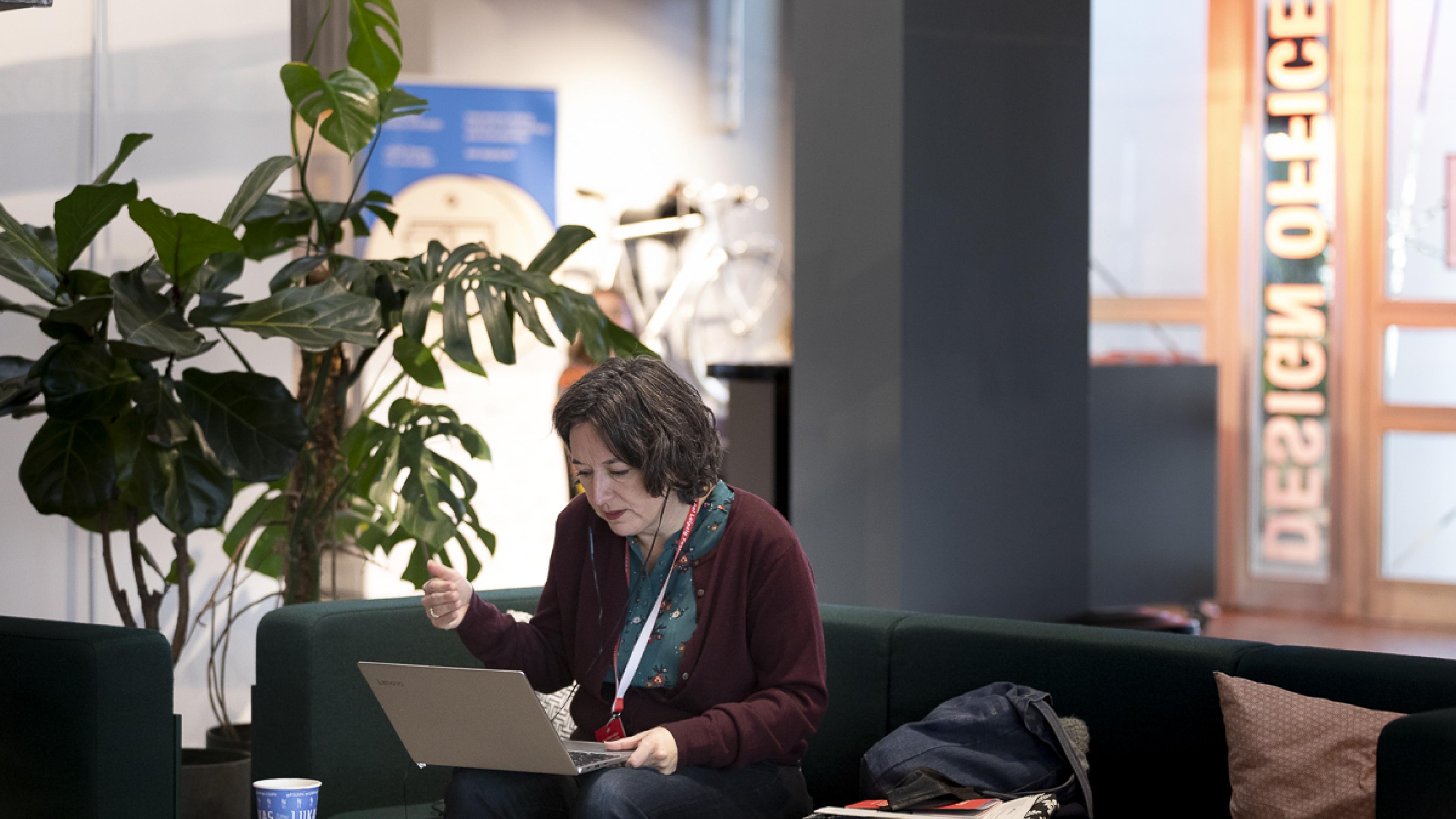 A woman with shoulder length dark hair is sitting on a couch in an office lounge. She put a laptop computer on her knees and is talking to the screen.