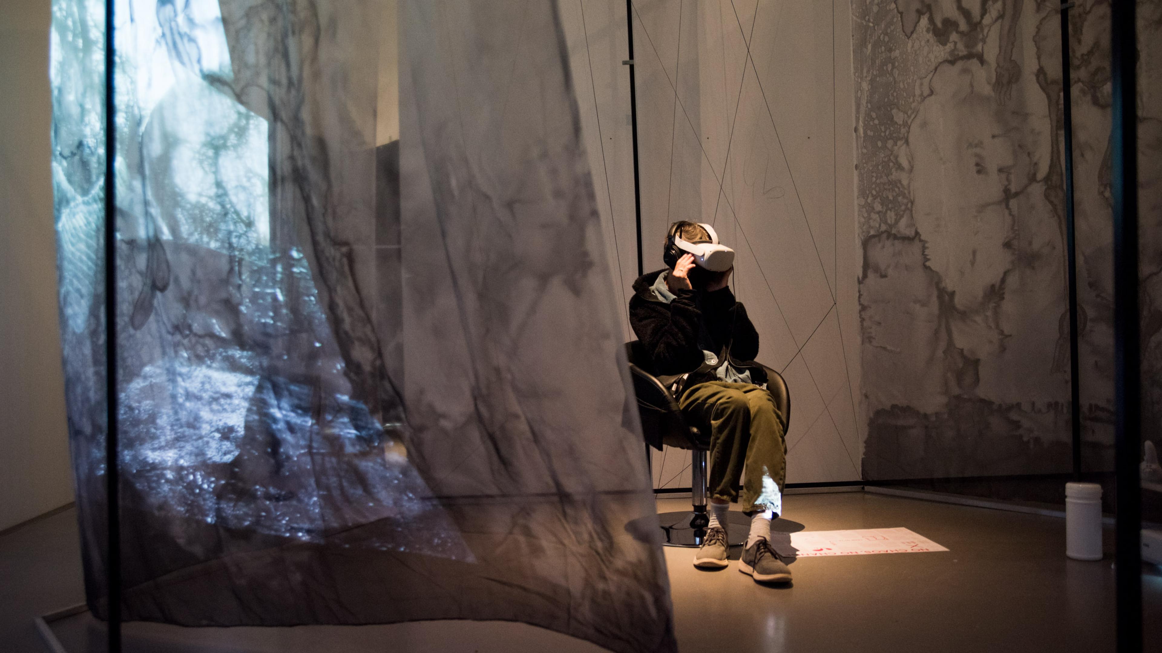 Ein Besucher von DOK Neuland trägt VR-Brille und große Kopfhörer. Er sitzt in einem Ledersessel, um ihn herum hängen halb-transparente graue Stoffbahnen von der Decke, auf denen eine Lichtprojektion sichtbar ist.
