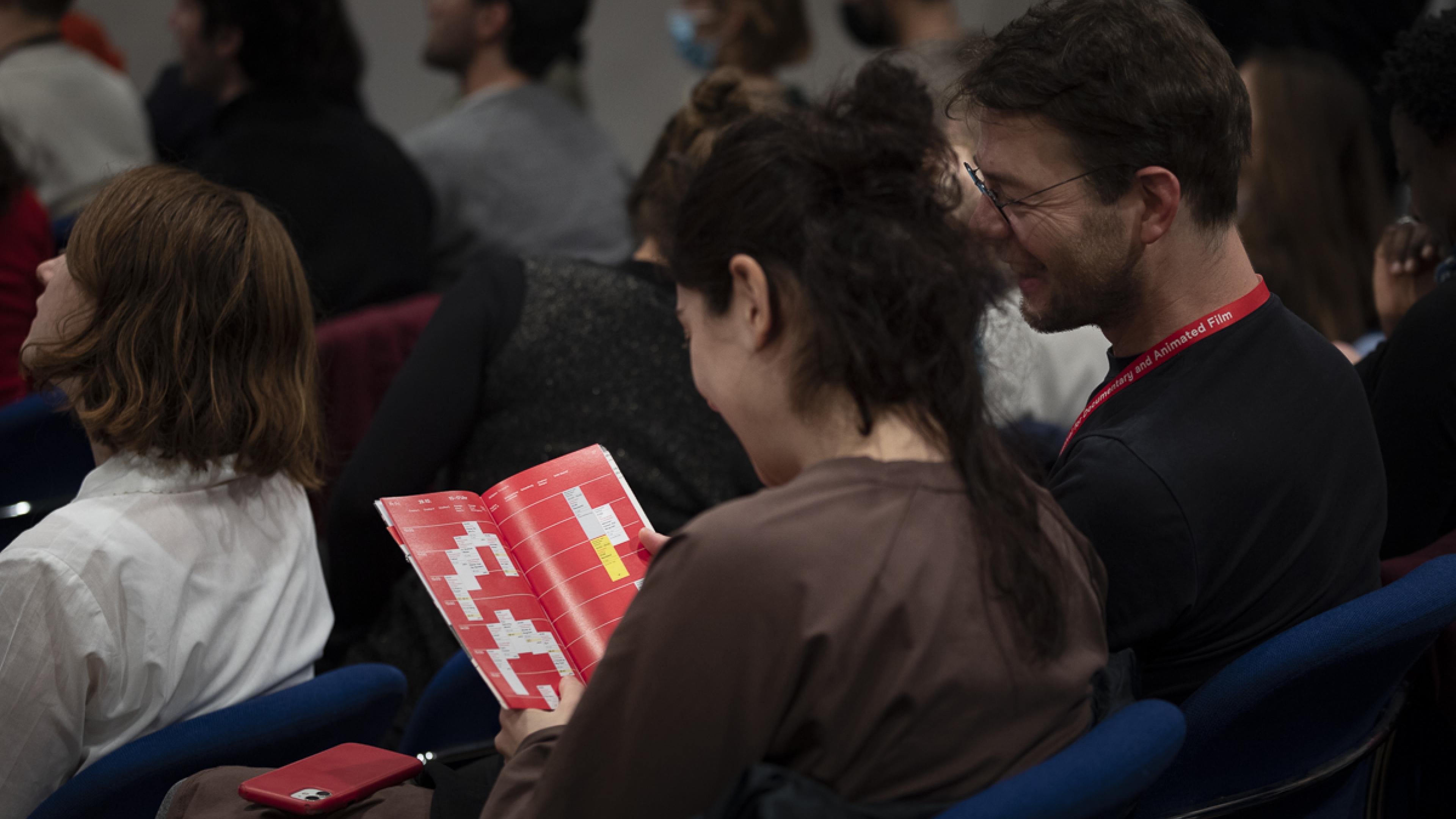 Zwei Kinobesucher*innen bei DOK Leipzig sitzen in einer Sitzreihe und beugen sich gemeinsam über das Timetable im Programmheft. Beide lächeln.