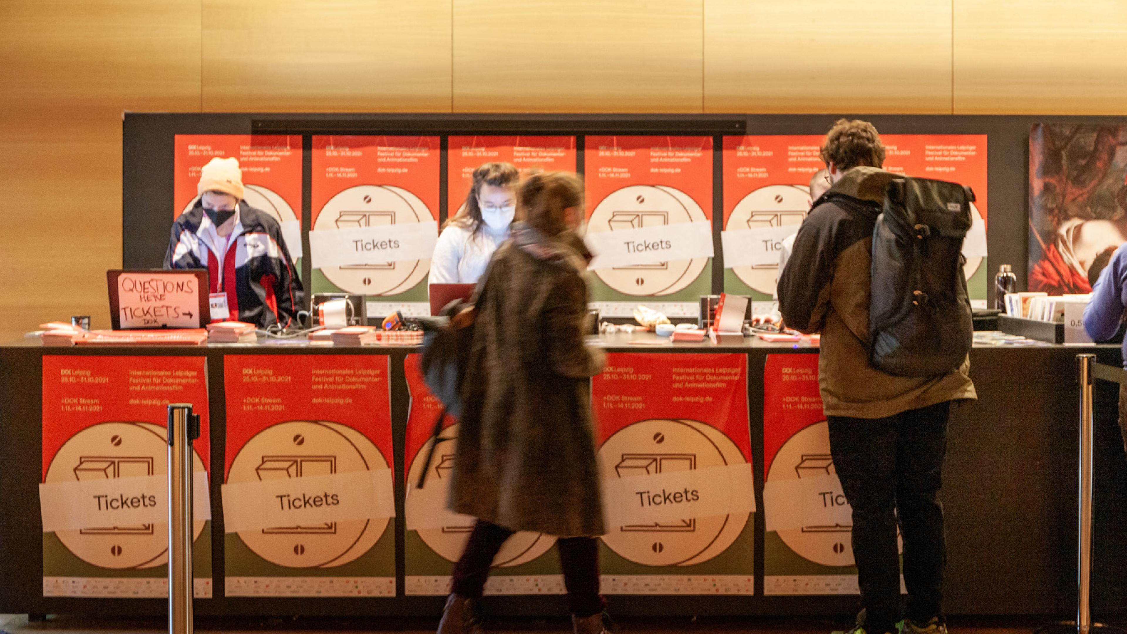 Der Tresen einer DOK-Ticketkasse. Der Tresen und die Wand dahinter sind mit zahlreichen DOK-Plakaten dekoriert. Zwei Festivalbesucher*innen kaufen gerade an zwei parallelen Schaltern Tickets.