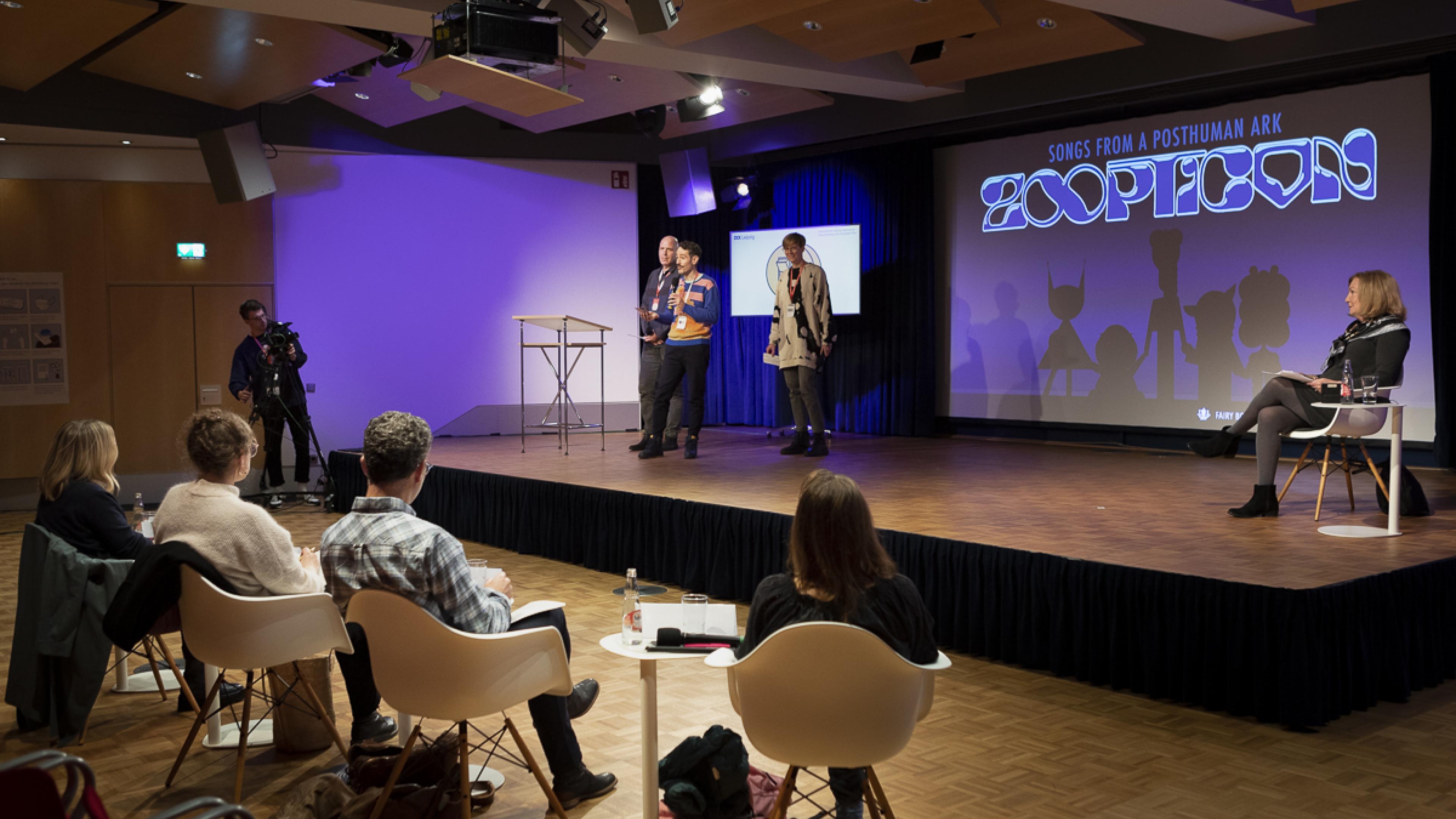 A pitch at DOK Industry: Three persons present on stage in front of a four-person panel. Moderator Elisabeth Klinck sits at the left side of the stage while listening.