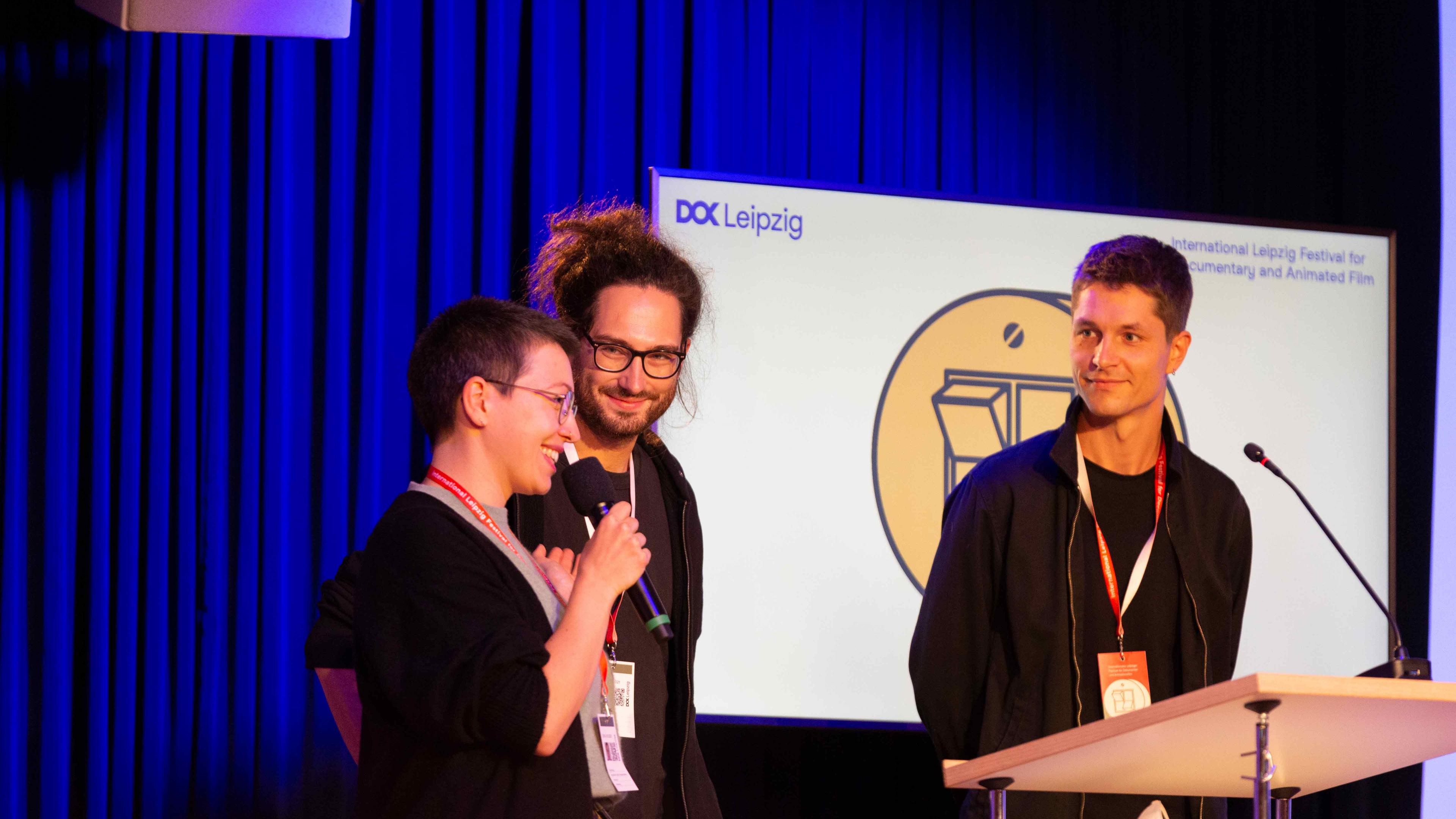 Three DOK Preview participants are standing behind a lectern on stage. The woman (left) speaks into a microphone, the two others (both male) are looking at her. All three are smiling.