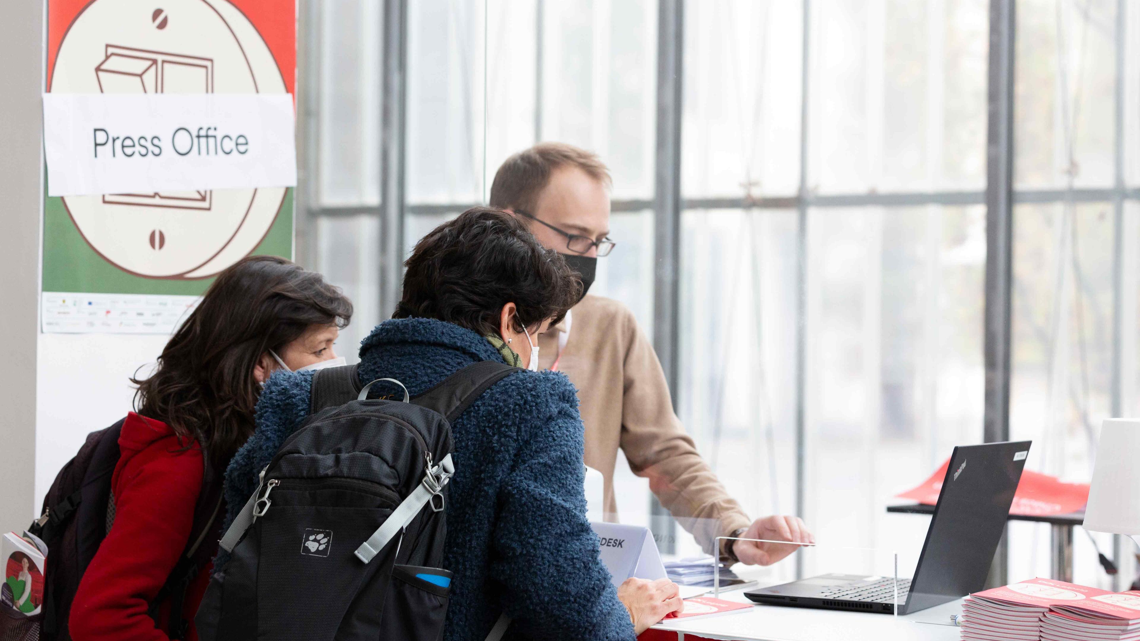 An einem Tresen im Festivalcenter stehen drei Personen und beugen sich gemeinsam über einen Laptop-Bildschirm, auf den sie konzentriert schauen. Alle tragen Mund-Nasenschutz. Um sie herum hängen DOK-Plakate.