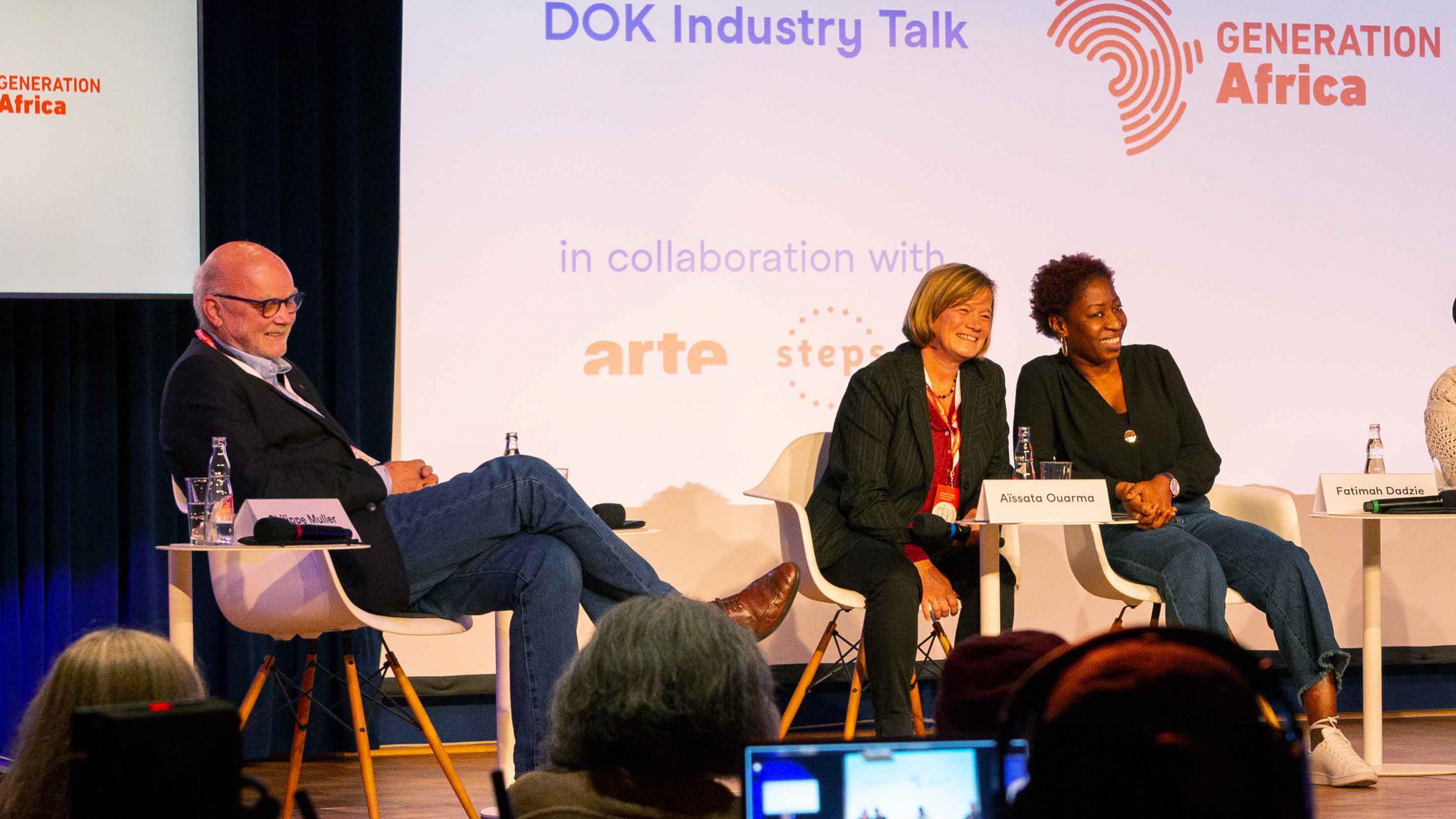 Three persons on stage of a panel, all of them laughing and looking to the audience. FLTR: A man in blue jeans, a woman with blond hair who is wearing a black suit, and a black woman with short hair. In the background a big screen with information on the event: DOK Industry Talk, below the logos of arte and steps.