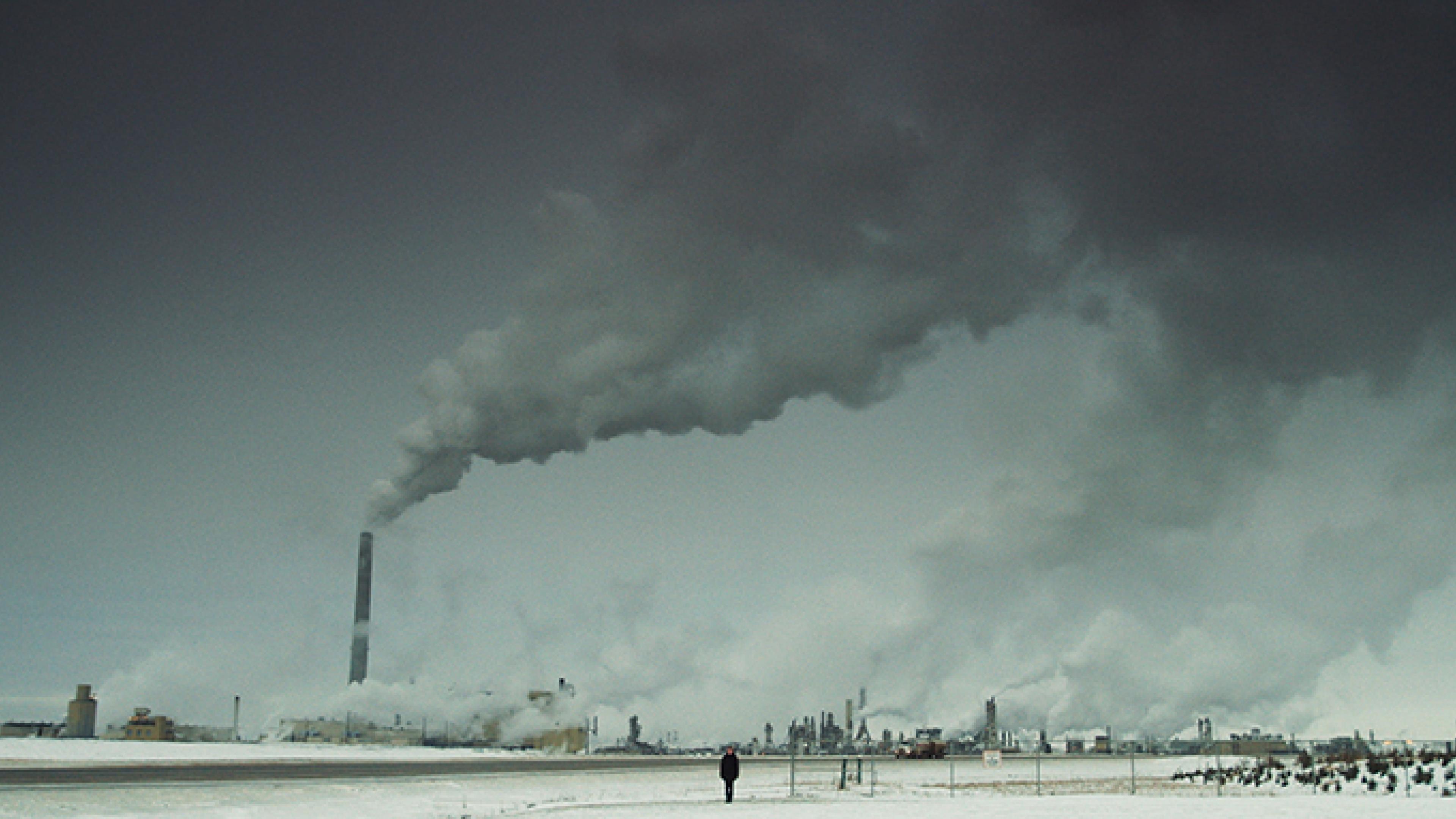 Eine wüstenähnliche, karge Ebene. Am Horizont sind viele rauchende Schornsteine mehrerer Raffinerien zu sehen. Mitten in dieser Szenerie steht eine schwarz gekleidete Person. 