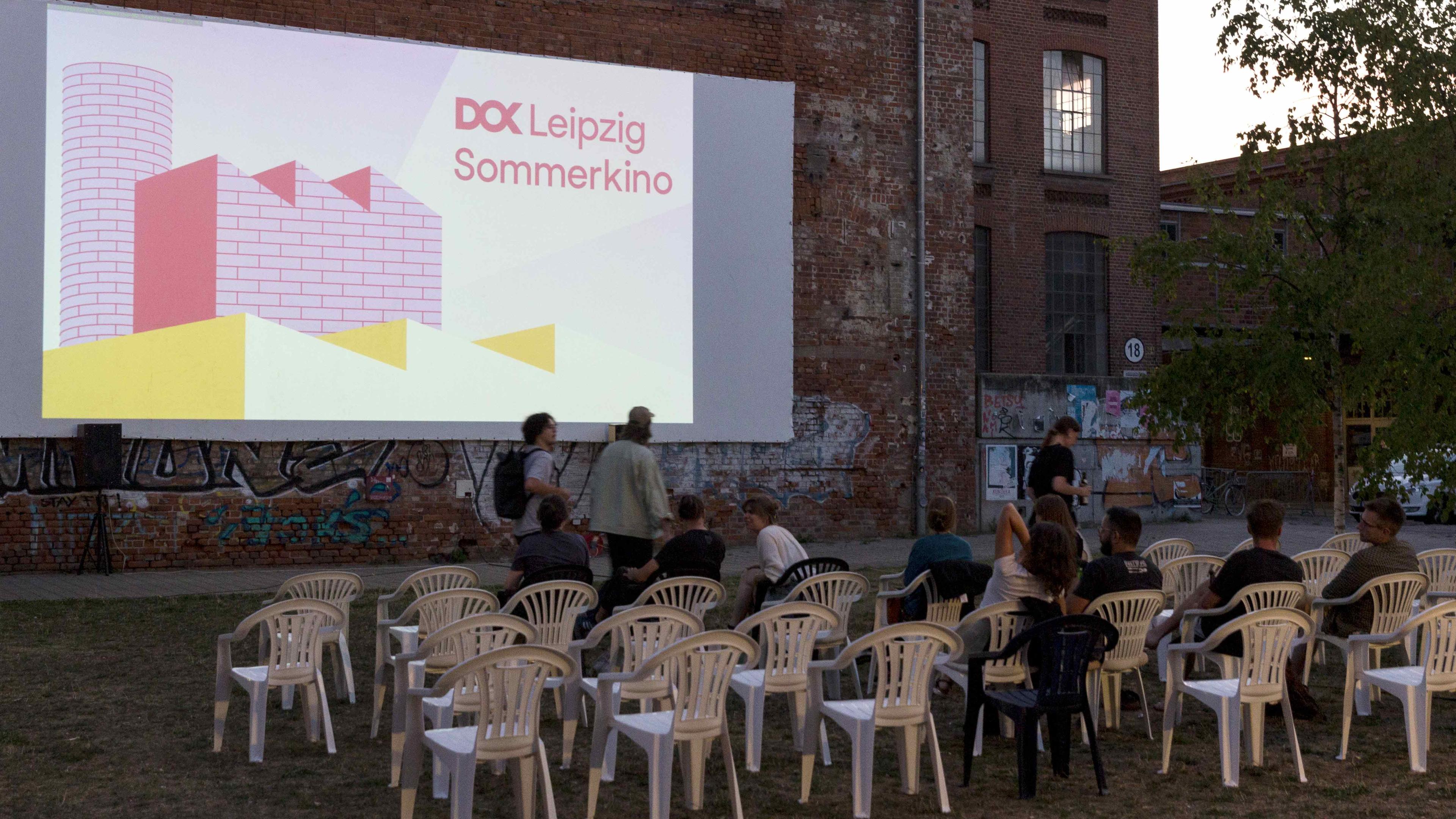 Auf einer Wiese stehen viele Plastiksessel in mehreren Reihen vor einer Leinwand. Auf der Leinwand steht "DOK Leipzig Sommerkino". Einige Stühle sind schon besetzt, vier Personen gehen vor den Stuhlreihen lang. Im Hintergrund die Abenddämmerung.