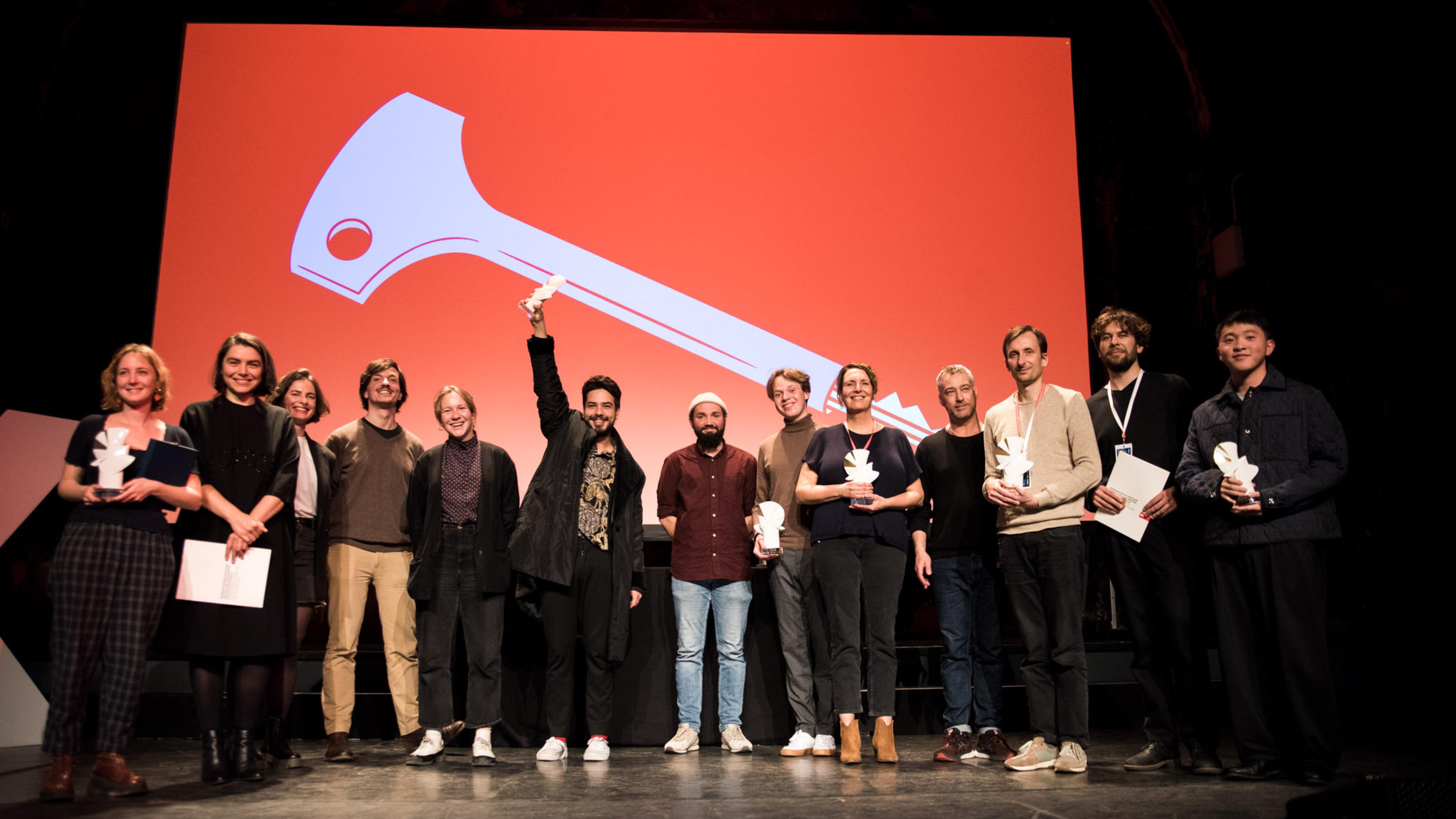 Eine Gruppe von 13 Personen steht auf einer Bühne. Alle blicken in Richtung Publikum und lächeln. Einige halten kleine Taubenfiguren aus weißem Porzellan in den Händen.