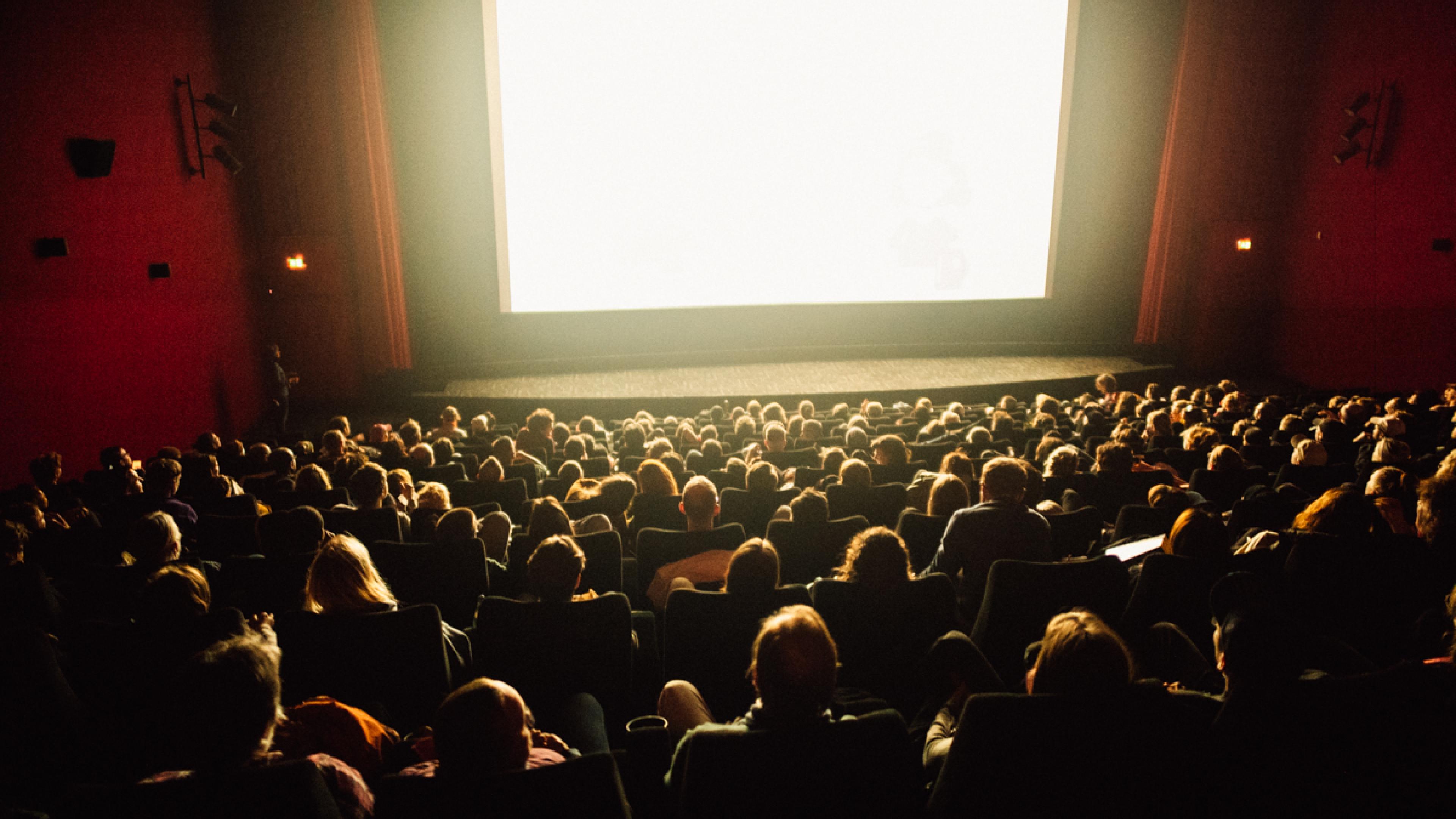 Ein vollbesetztes Kino bei einer Filmvorführung. Die Leinwand strahlt helles, weißes Licht über die Köpfe der Zuschauer*innen.