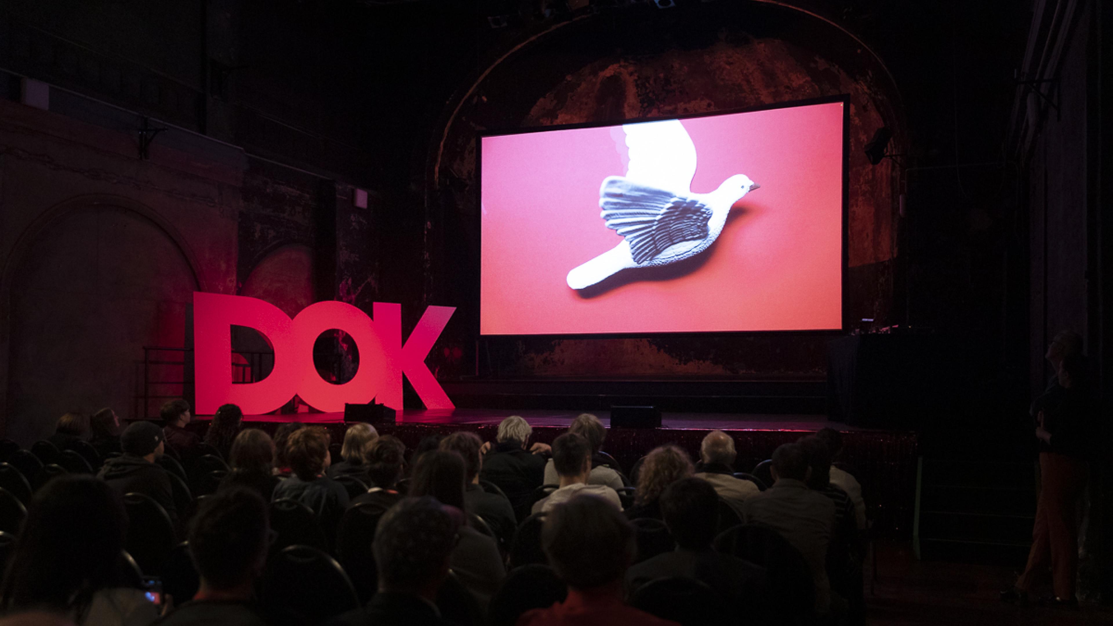A stage in a big hall. On stage three huge letters "D-O-K" and a big screen showing a white dove. 