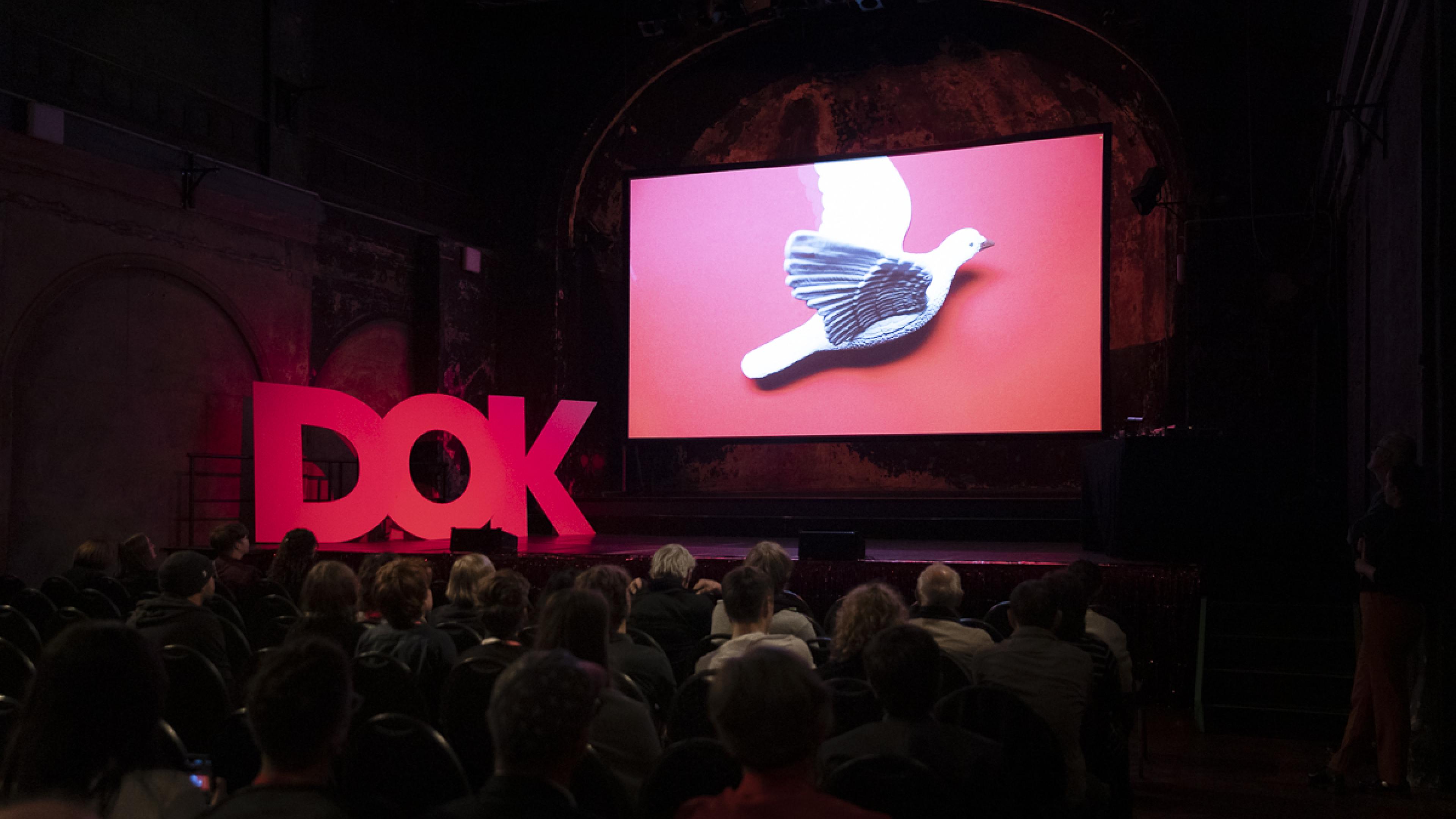 A stage in a big hall. On stage three huge letters "D-O-K" and a big screen showing a white dove.