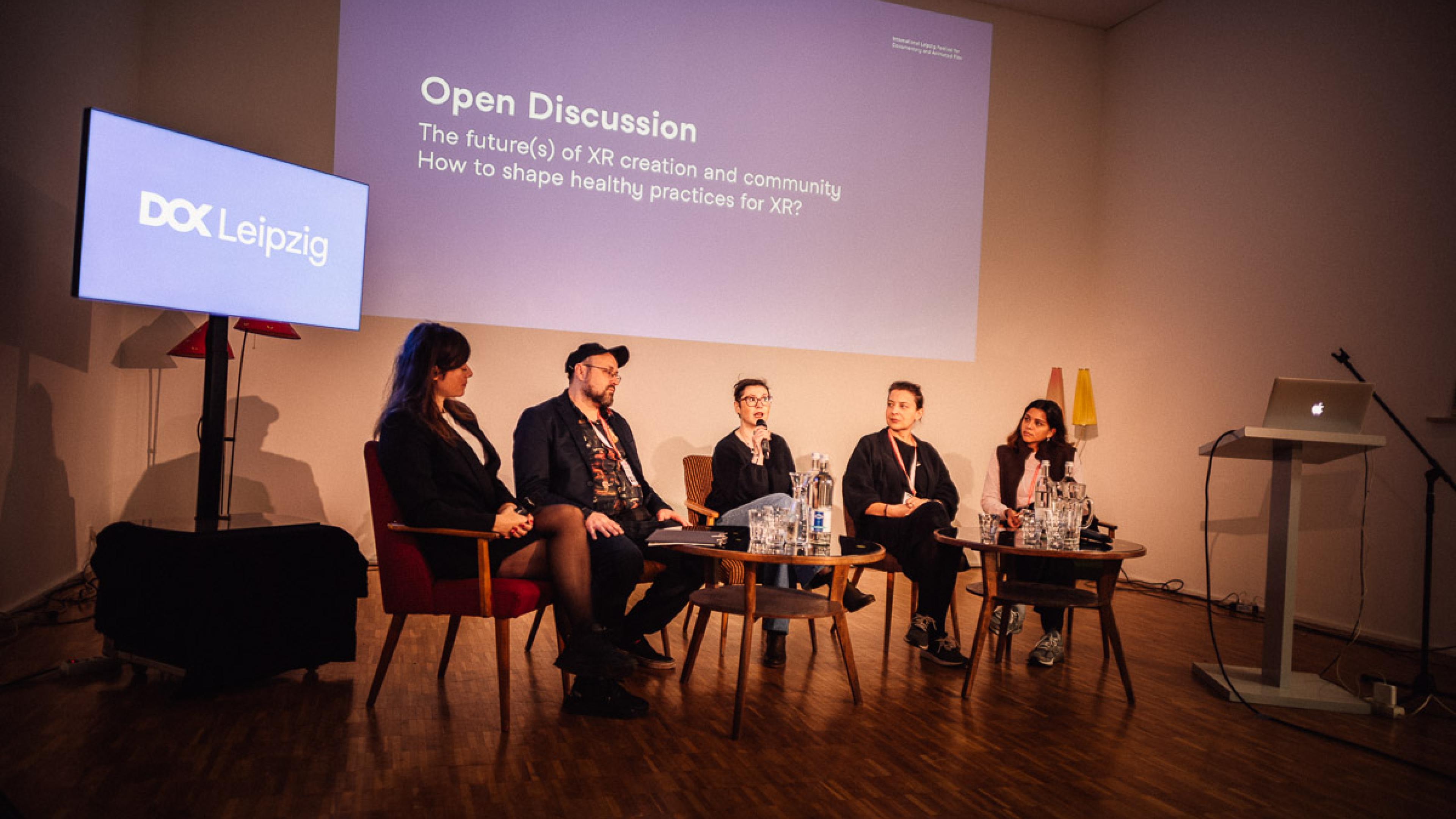 Five persons are sitting on sofa chairs on a discussion panel. The woman in the middle is talking into a microphone. In the background a blue info graphic is projected on the wall. It says: "Open discussion. The future(s) of XR creation and community."