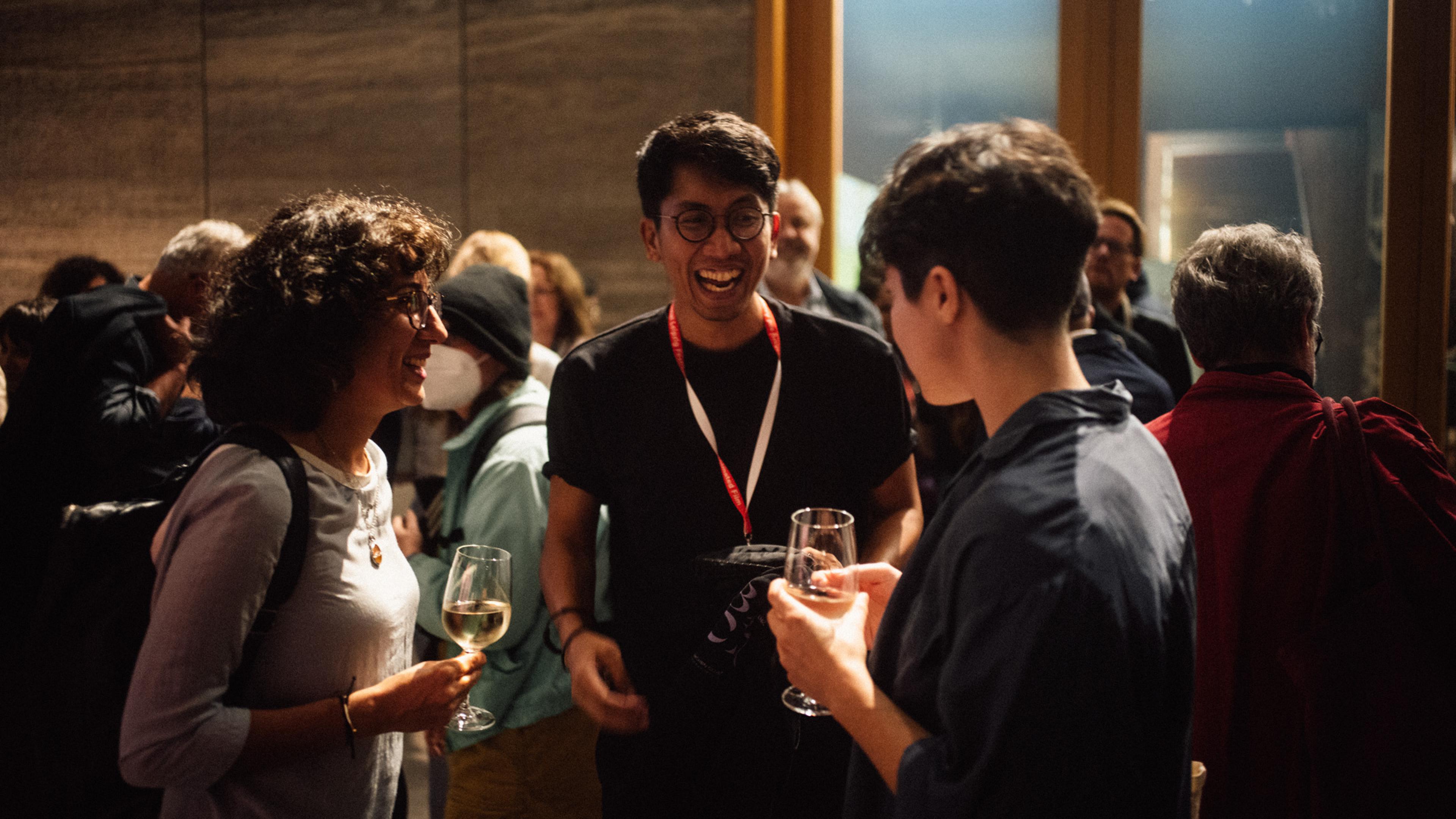 Three people are standing together and talking. They are holding gasses with drinks in their hands. The man in the middle laughs heartily. Other people are standing around the group, all engrossed in conversation.