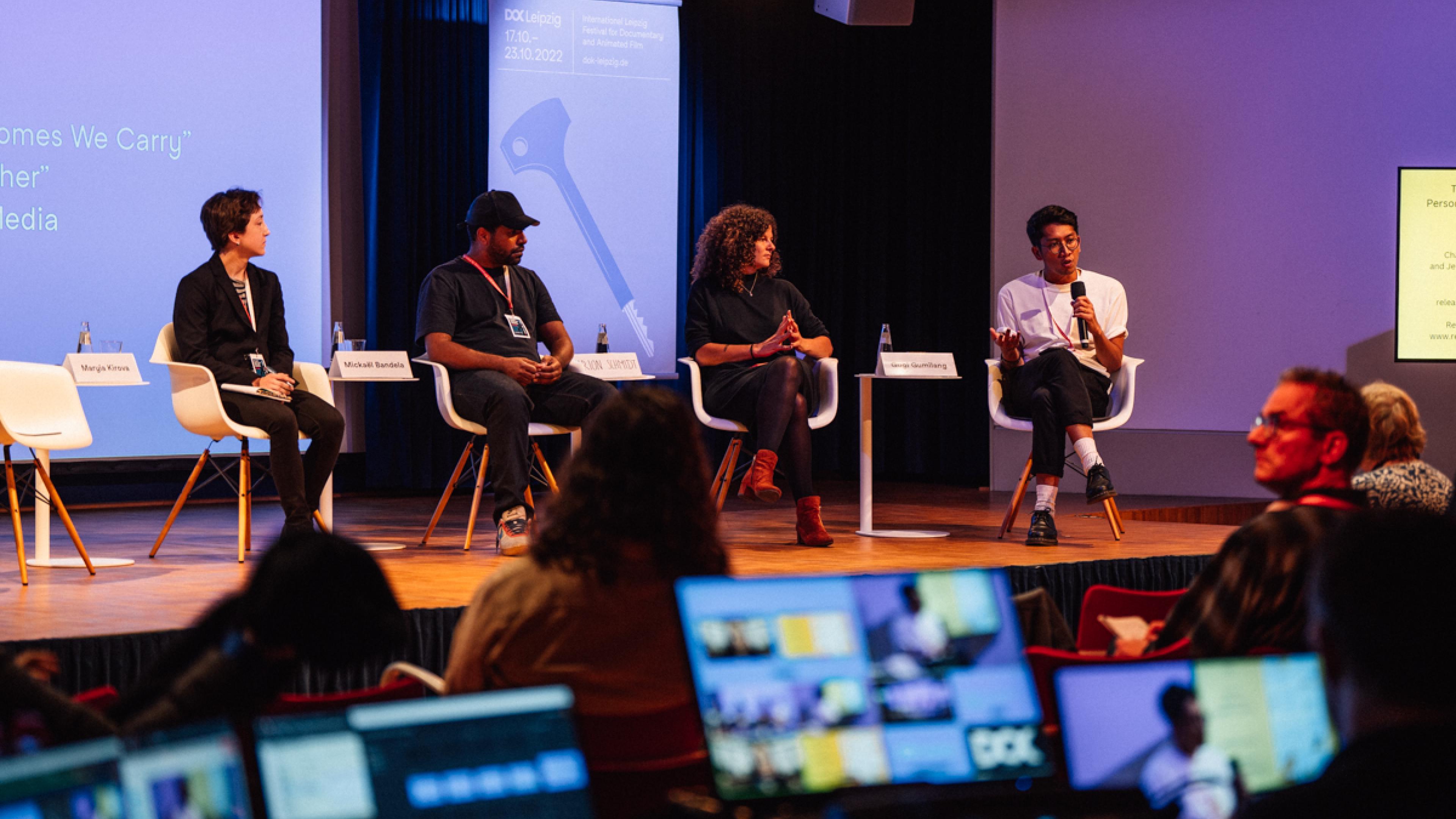Four persons (two women, two men) are sitting on a panel. The man at the right is talking into a handheld microphone, the others are listening. In the front, the monitors of a live-editing suite light up in the dark.