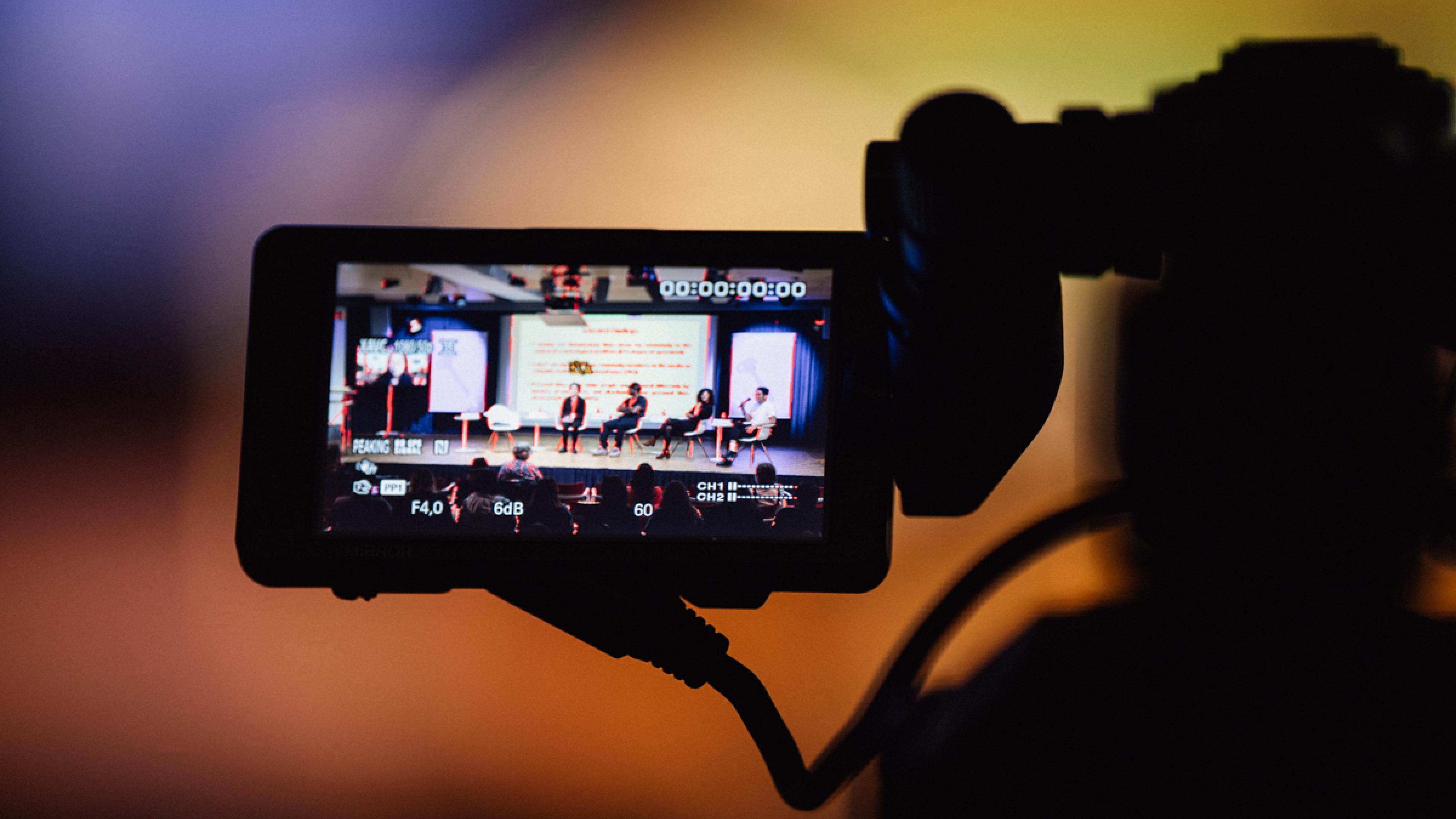 The monitor of a TV camera. On the screen the setting of a panel with four persons sitting on stage, one chair (left) is empty.
