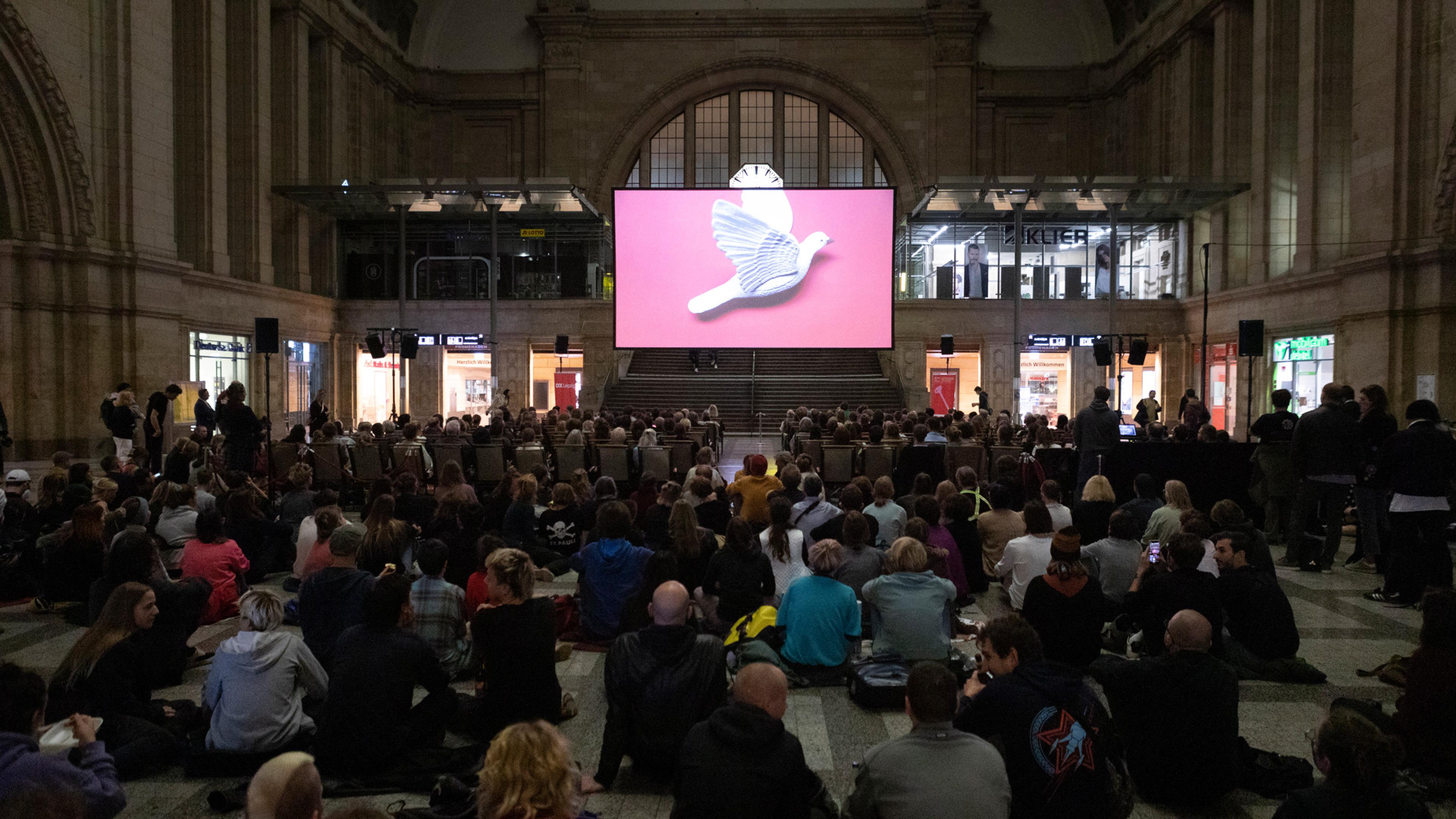 In einer großen Halle ist eine Kinoleinwand mit Sitzreihen aufgebaut. Die Sitzreihen sind voll besetzt, rundherum und dahinter sitzen zahlreiche Zuschauer*innen auf dem Boden. Auf der Leinwand fliegt eine weiße Taube auf pinkem Hintergrund.