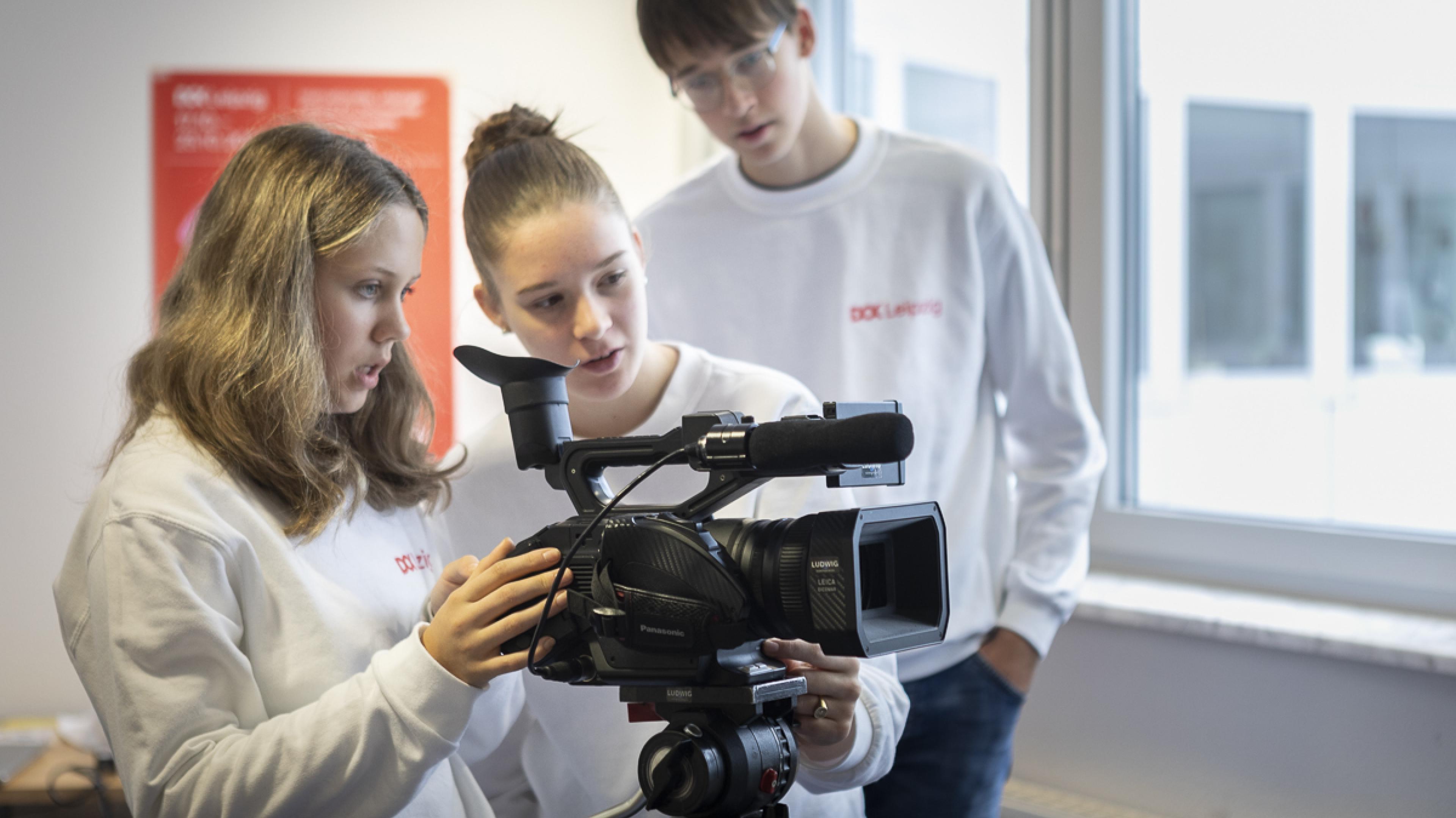 Drei Jugendliche – zwei Mädchen und ein Junge – beugen sich über eine Videokamera auf einem Stativ. 