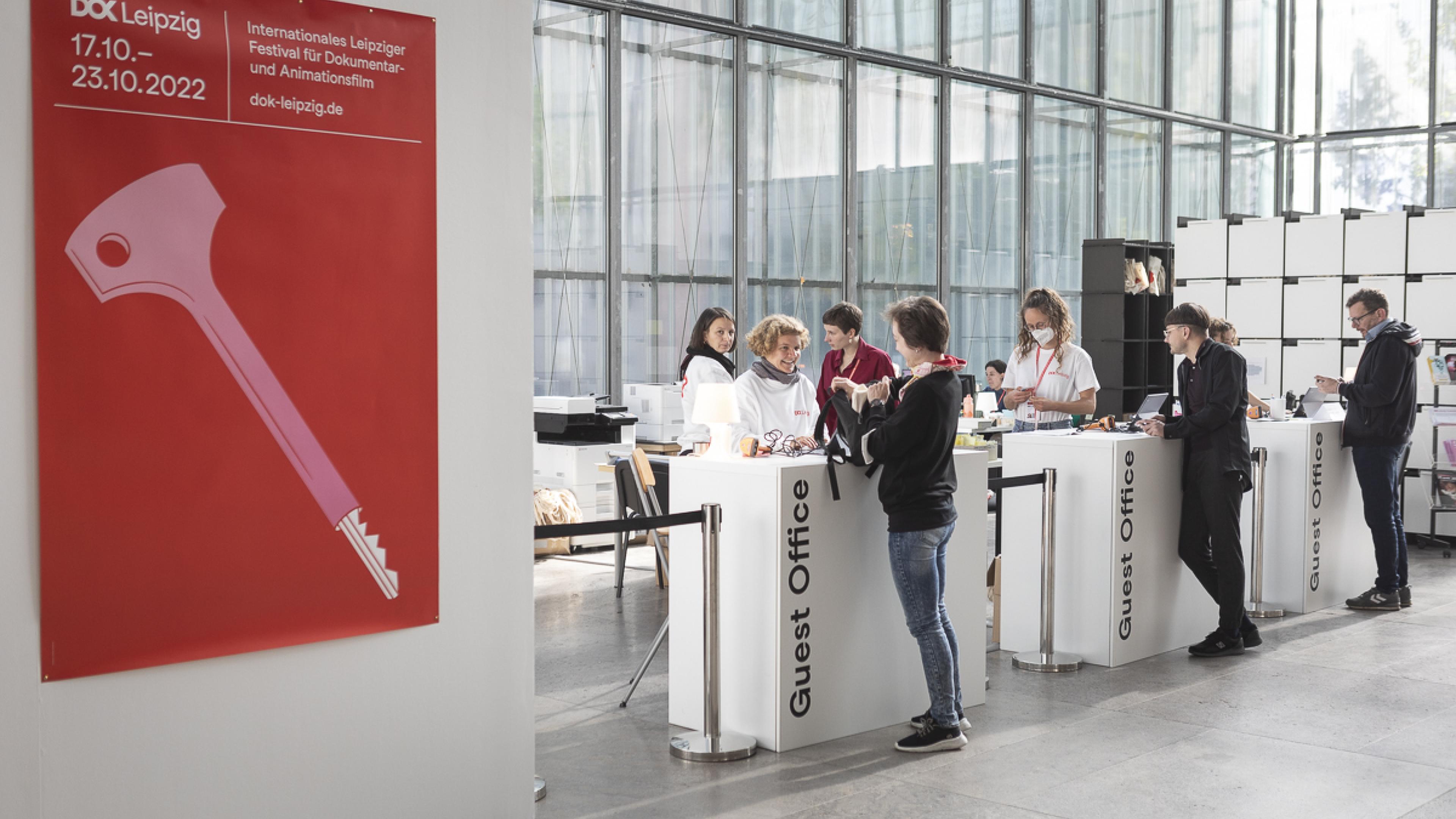 Three white reception counters at the festival centre: At each counter a team member is talking to a festival visitor. At the left the festival poster is hanging at a huge white wall.
