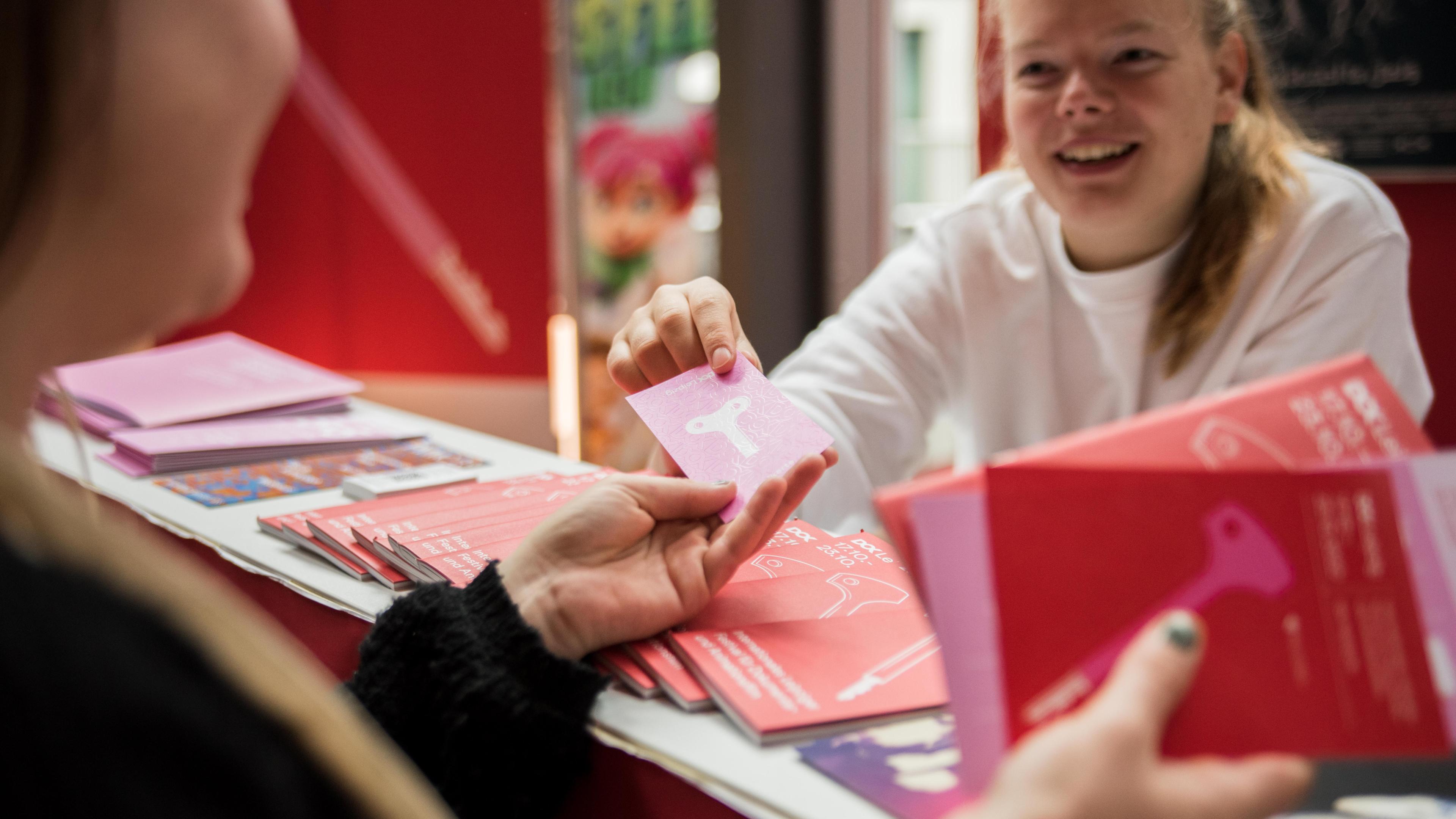 Eine Frau überreicht einer anderen Frau ein Kinoticket. Die Empfängerin hält ein Programmheft von Dok Leipzig in der Hand.