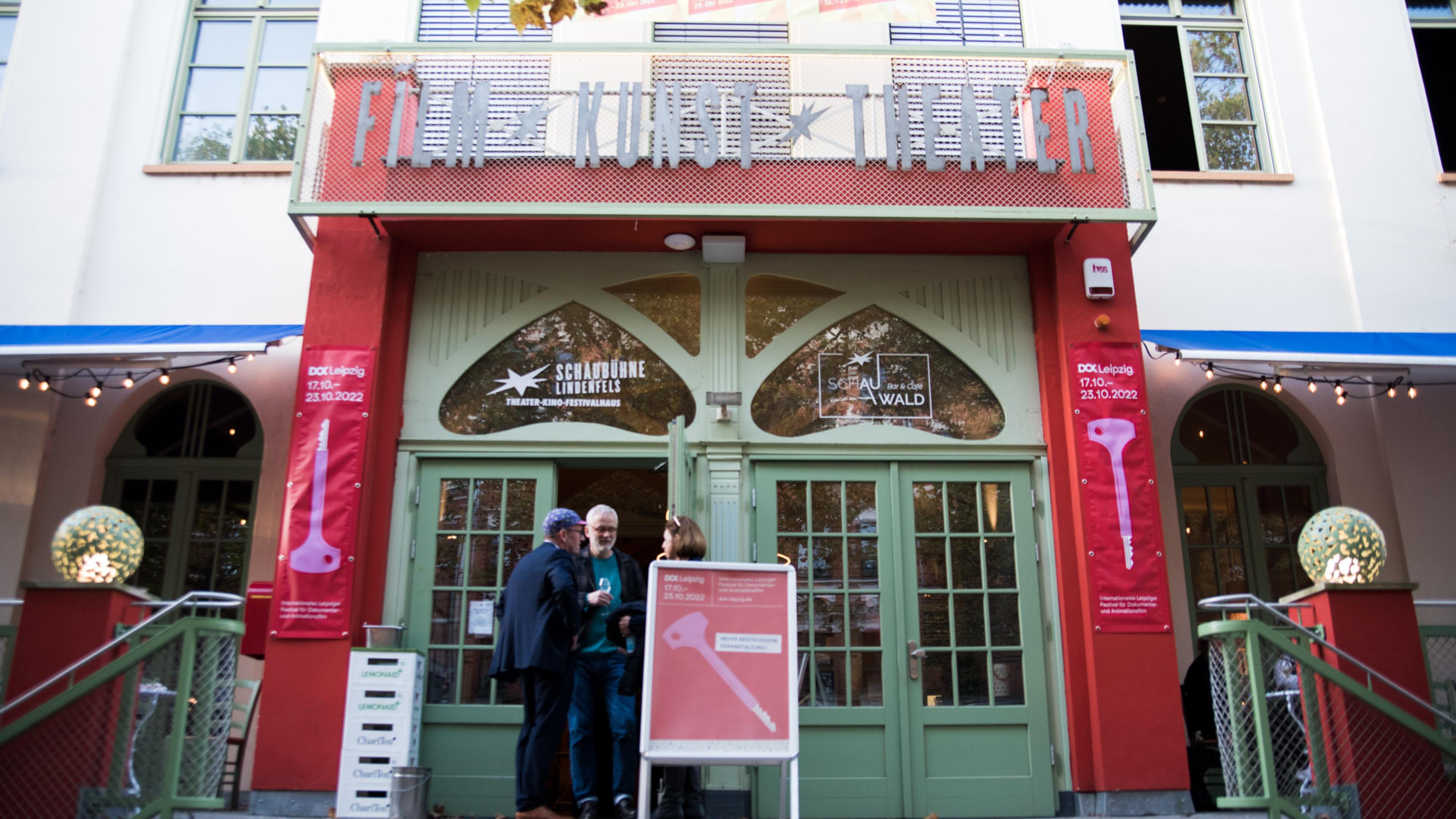 Der Eingang zum Festivalkino Schaubühne Lindenfels. Drei Personen stehen im Gespräch vor der Tür. Links und rechts rahmen DOK Leipzig Banner den Eingang, in der Mitte steht ein Aussteller mit dem Festivalplakat.