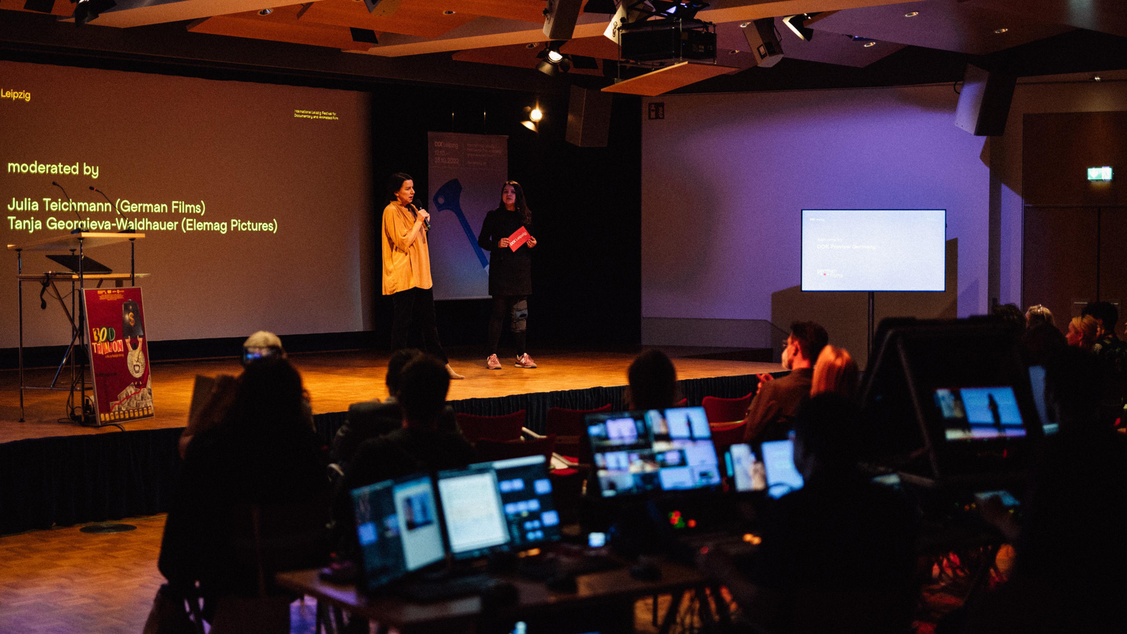 Two women are presenting on a stage in a conference room. In the middle of the audience is a live editing suite with several monitors. The monitors light up. 