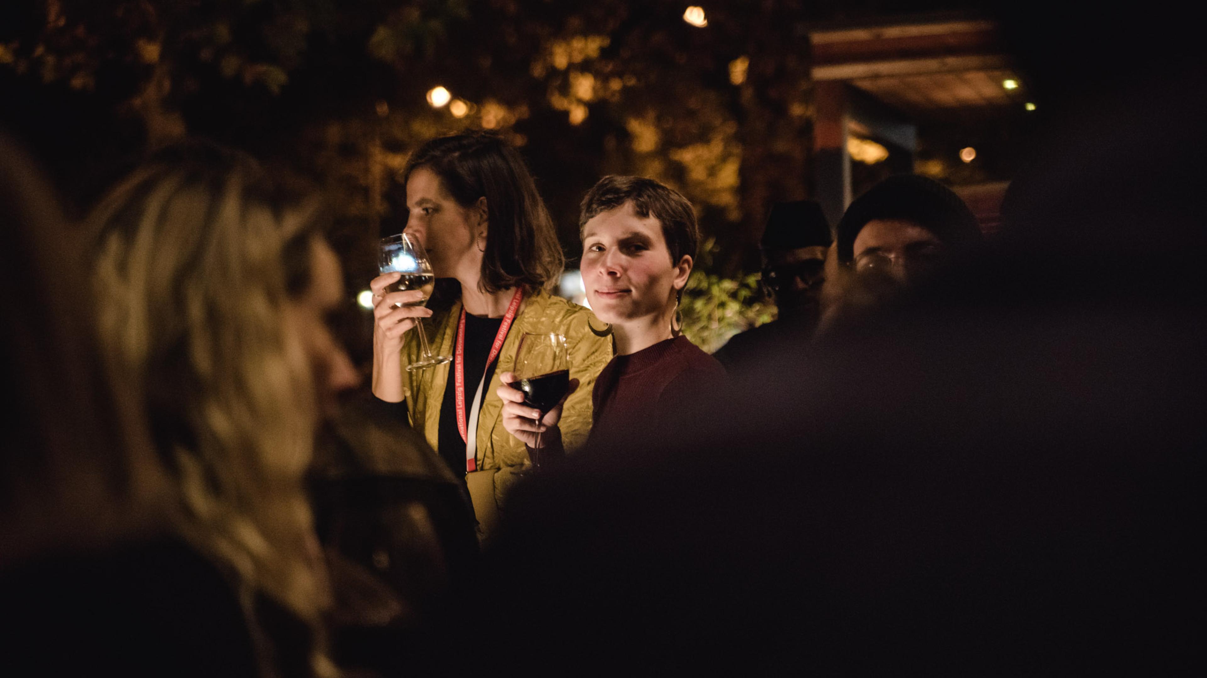 Bei einer Abendveranstaltung steht eine Frau mit kurzen Haaren und einem Glas Rotwein in der Hand umringt von weiteren Personen. Sie blickt seitlich über ihre Schulter direkt in die Kamera und lächelt.