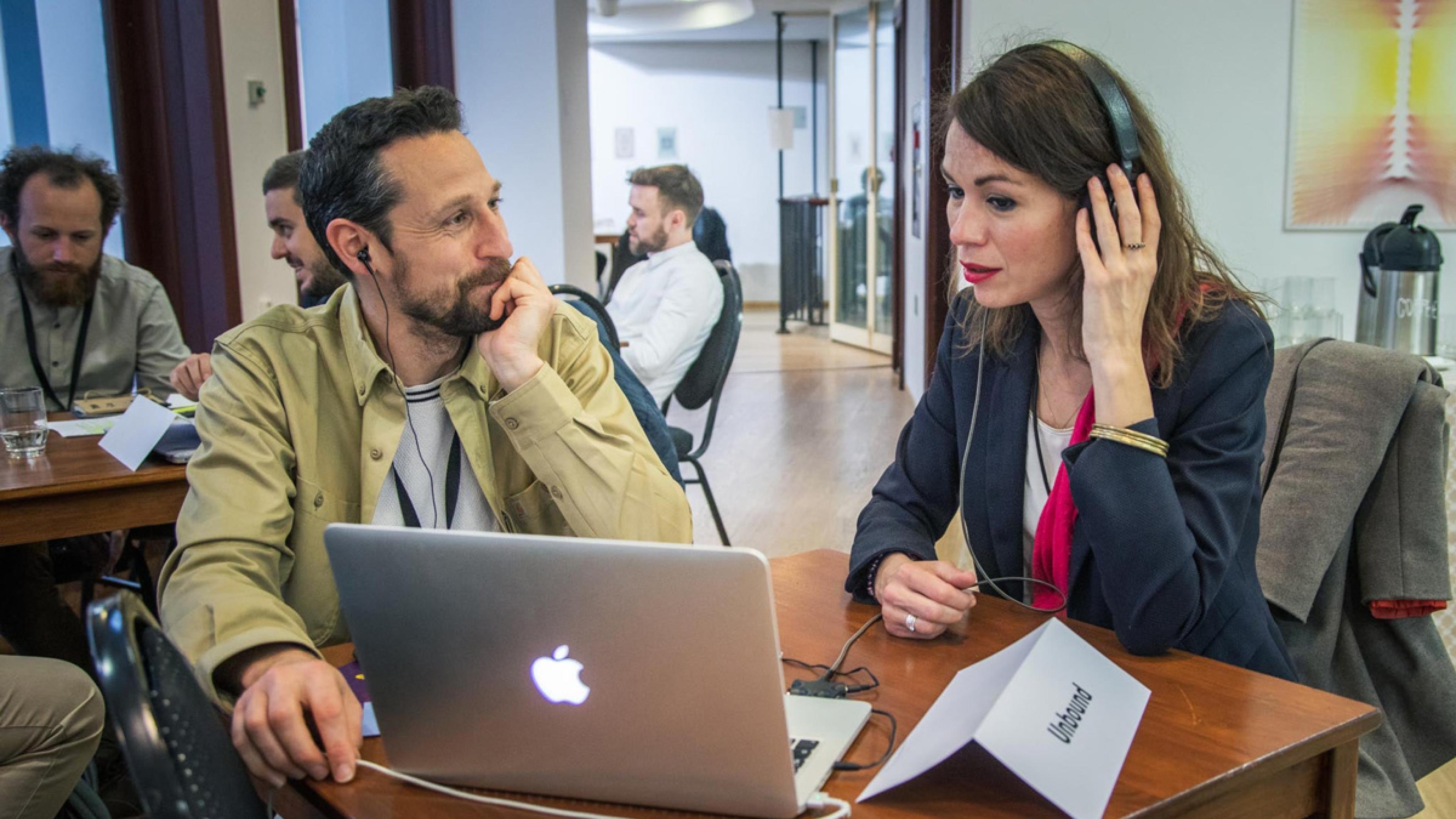 Two persons sit at a small table, the woman (at the right) wears earphones and looks at a laptop. The man (at the left) watches her.